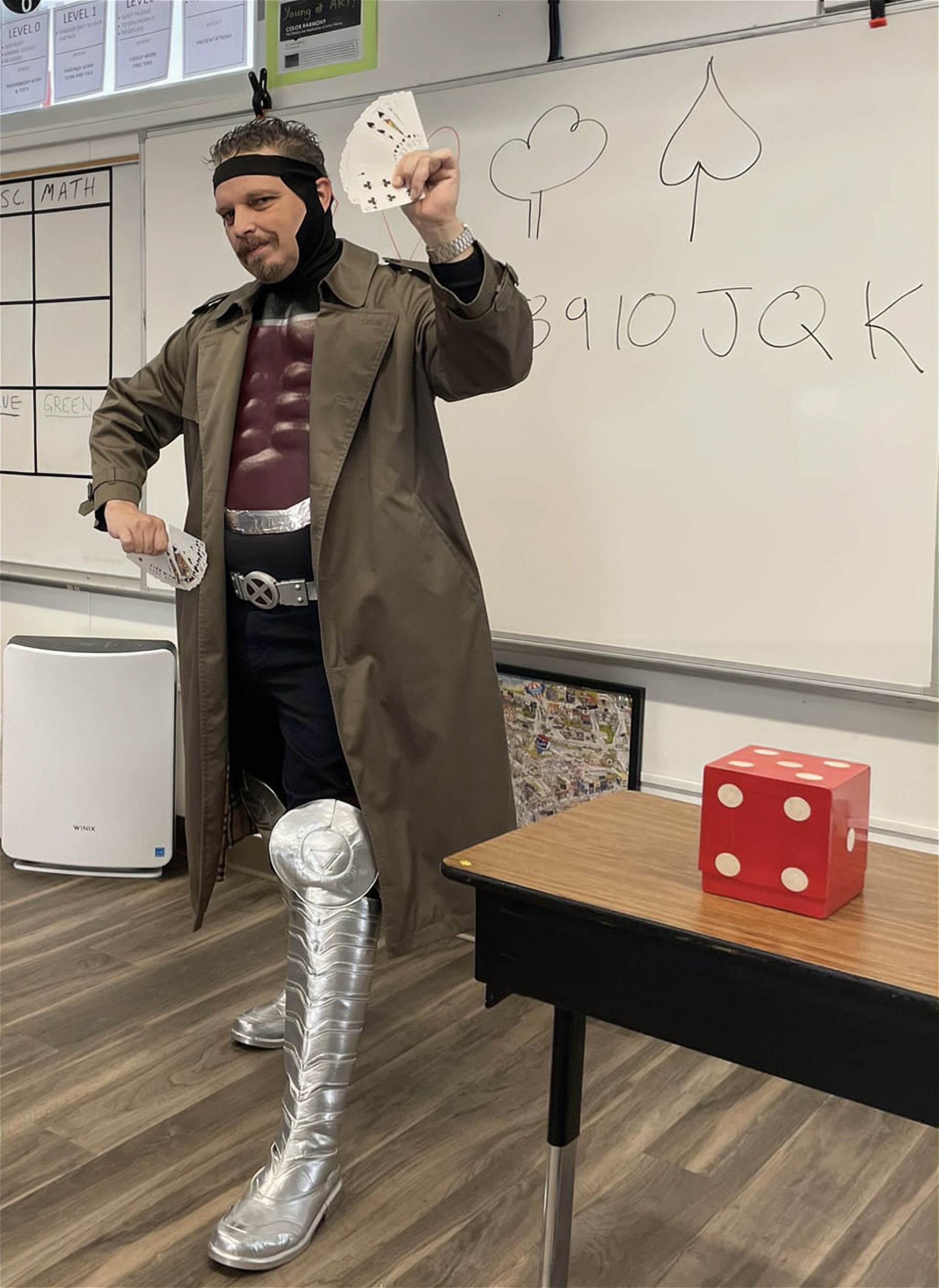 Person dressed as a comic book character, wearing a trench coat, large silver boots, and a costume with a chest symbol, standing in a classroom with a whiteboard, a large red die on a table, and holding playing cards.