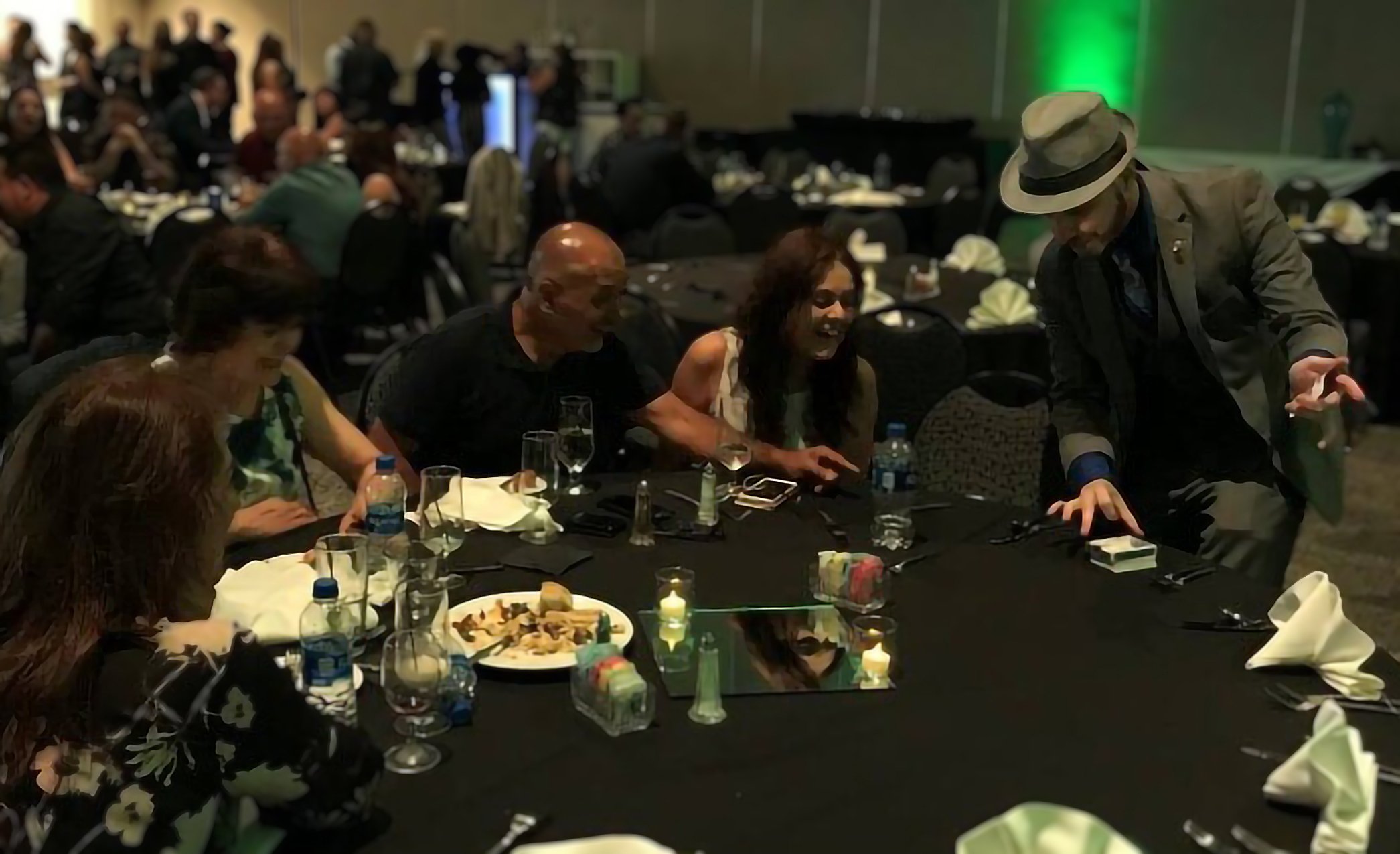 Several spectators at a Frito Lay corporate event point in astonishment as John reaches for a deck of cards on the table that wasn't there seconds ago. 