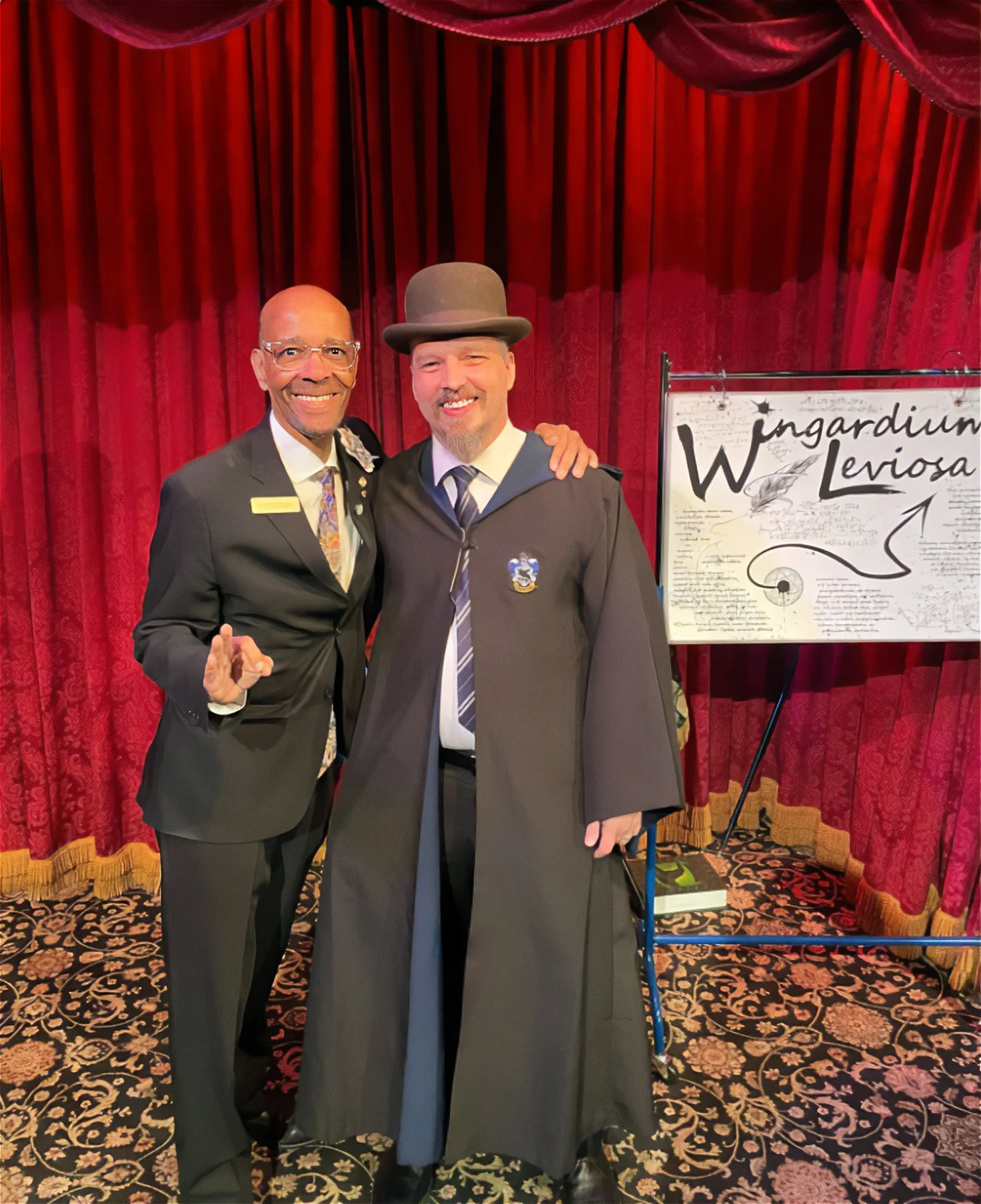 John clougherty standing with Jack Goldfinger in the Parlor of Prestidigitation at the Magic Castle. John is wearing a Ravenclaw robe with a custom Wingardium Leviosa art in the background in front of a Red Velvet stage curtain.