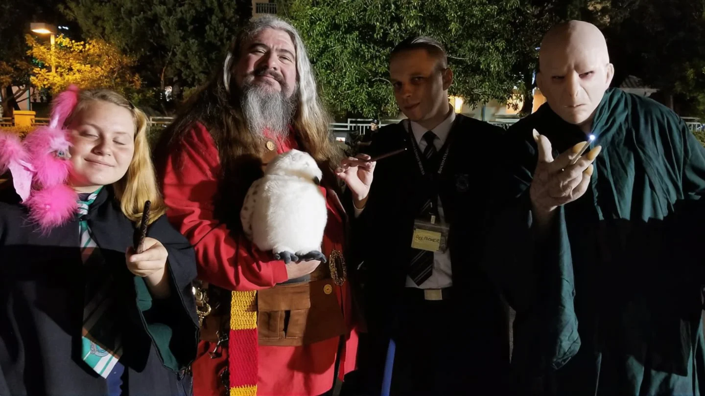 Group of four people dressed in costumes at a nighttime outdoor event, with trees and lights in the background. One person is dressed as a fantasy character, another as a girl with pink feathers, one as a wizard or Gandalf, and another as a creature 