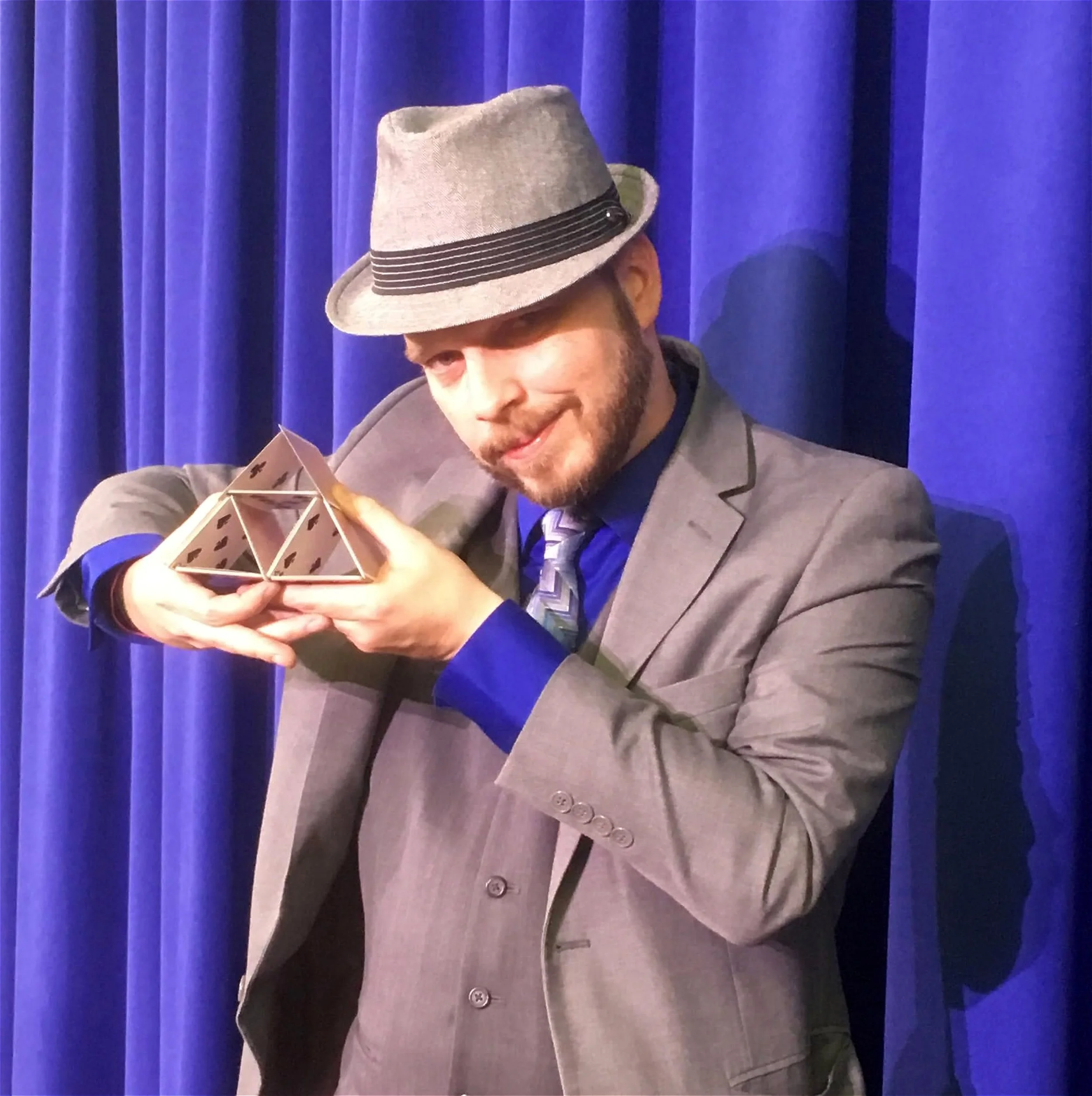 A man with a beard and mustache wearing a gray fedora hat, gray suit, blue shirt, and patterned tie holding a small wooden pyramid puzzle near his face in front of blue curtains.