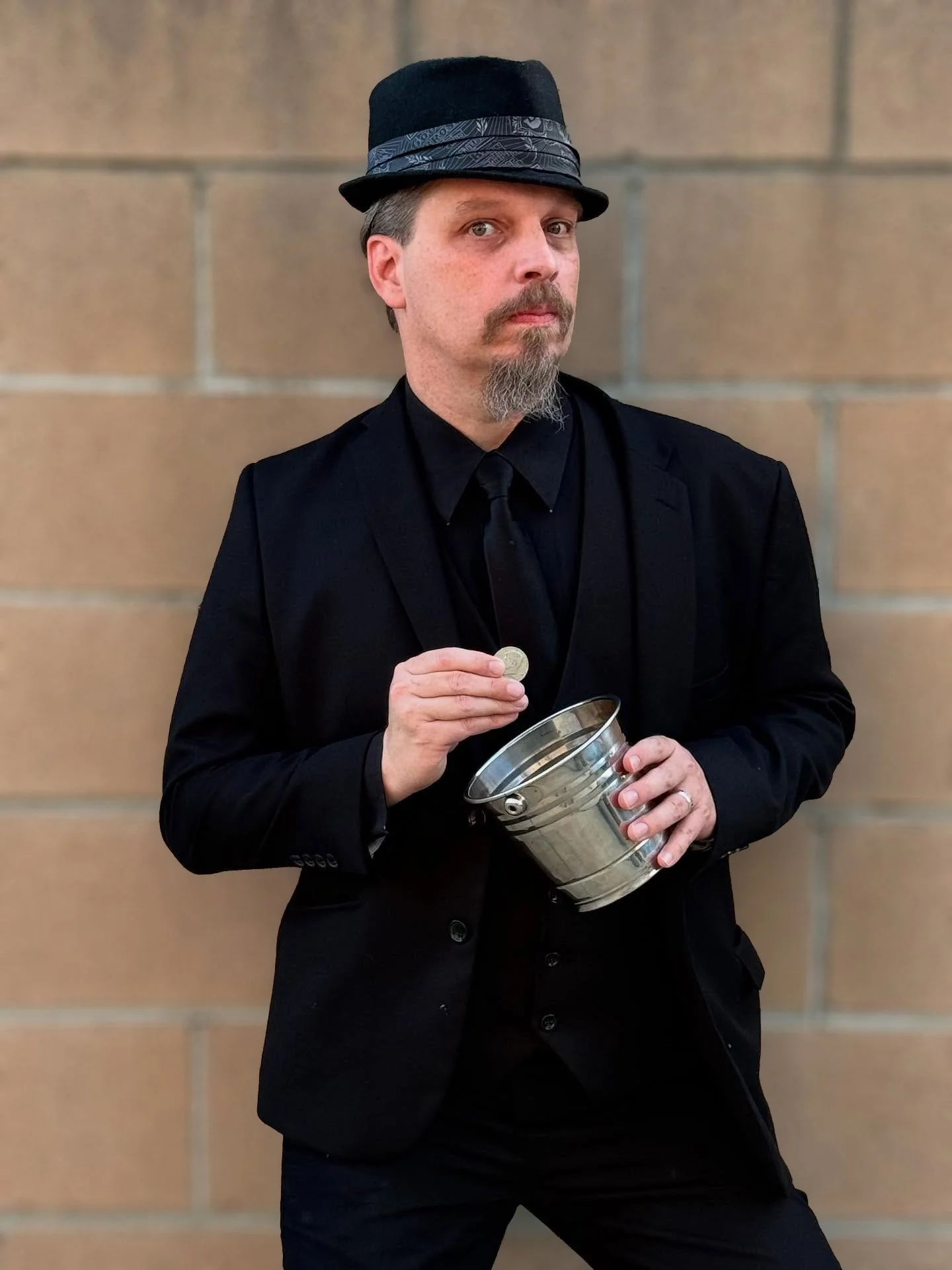 A man dressed in black, wearing a hat, holding a coin and a metal bucket, standing against a brick wall.