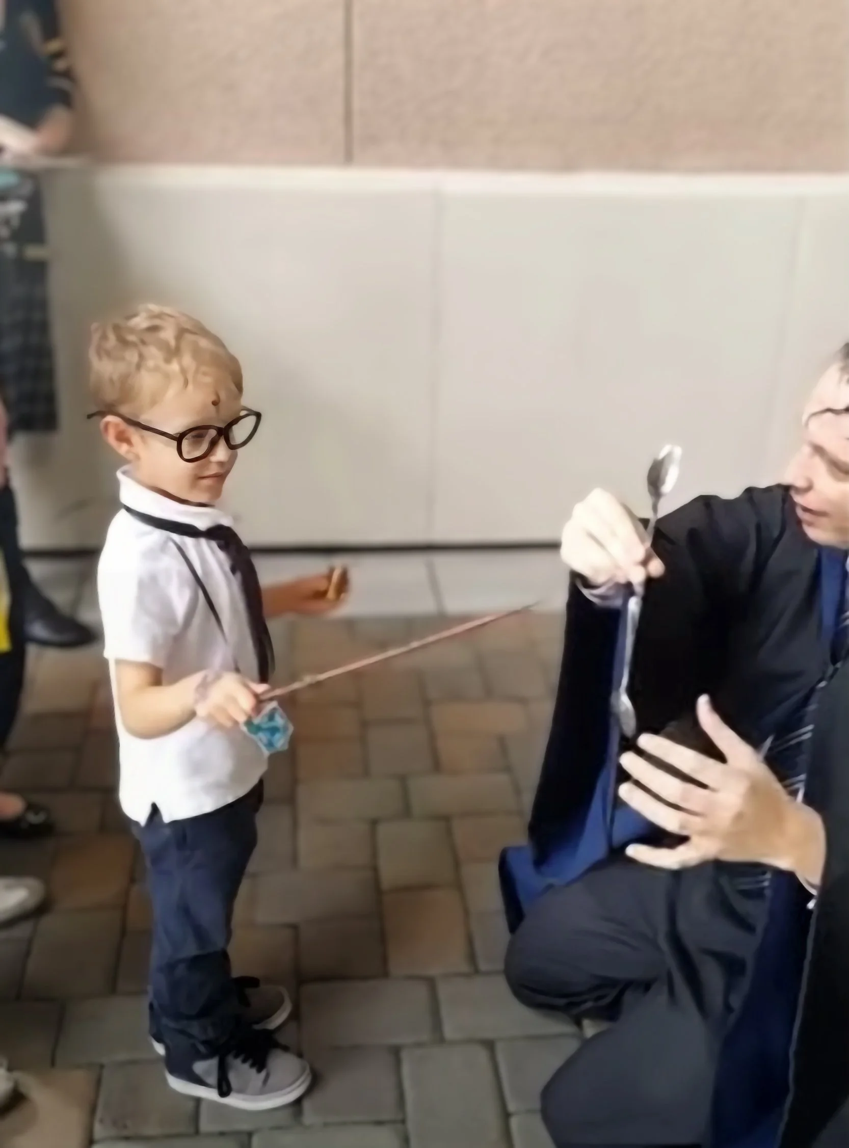 A young boy wearing glasses and a white shirt with a black tie stands in front of a man wearing a black robe, who is holding a silver spoon. The boy is holding a string attached to the spoon, and the boy has a small blue face mask hanging from his sh