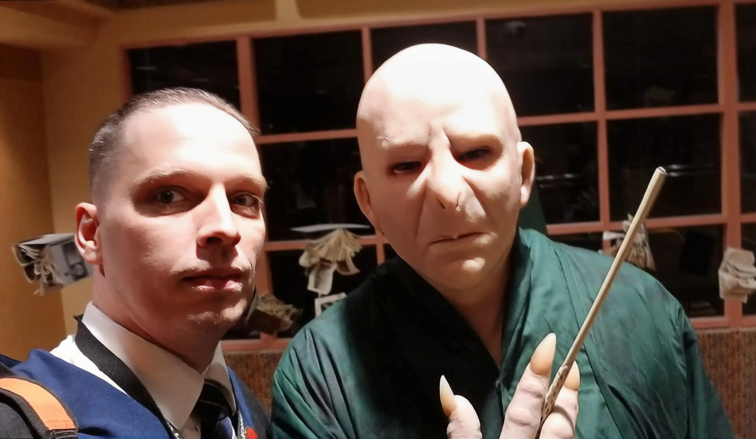 Two men posing for a photo indoors, one with short hair in a school uniform, and the other with a bald head and a grim expression, holding a chopstick.