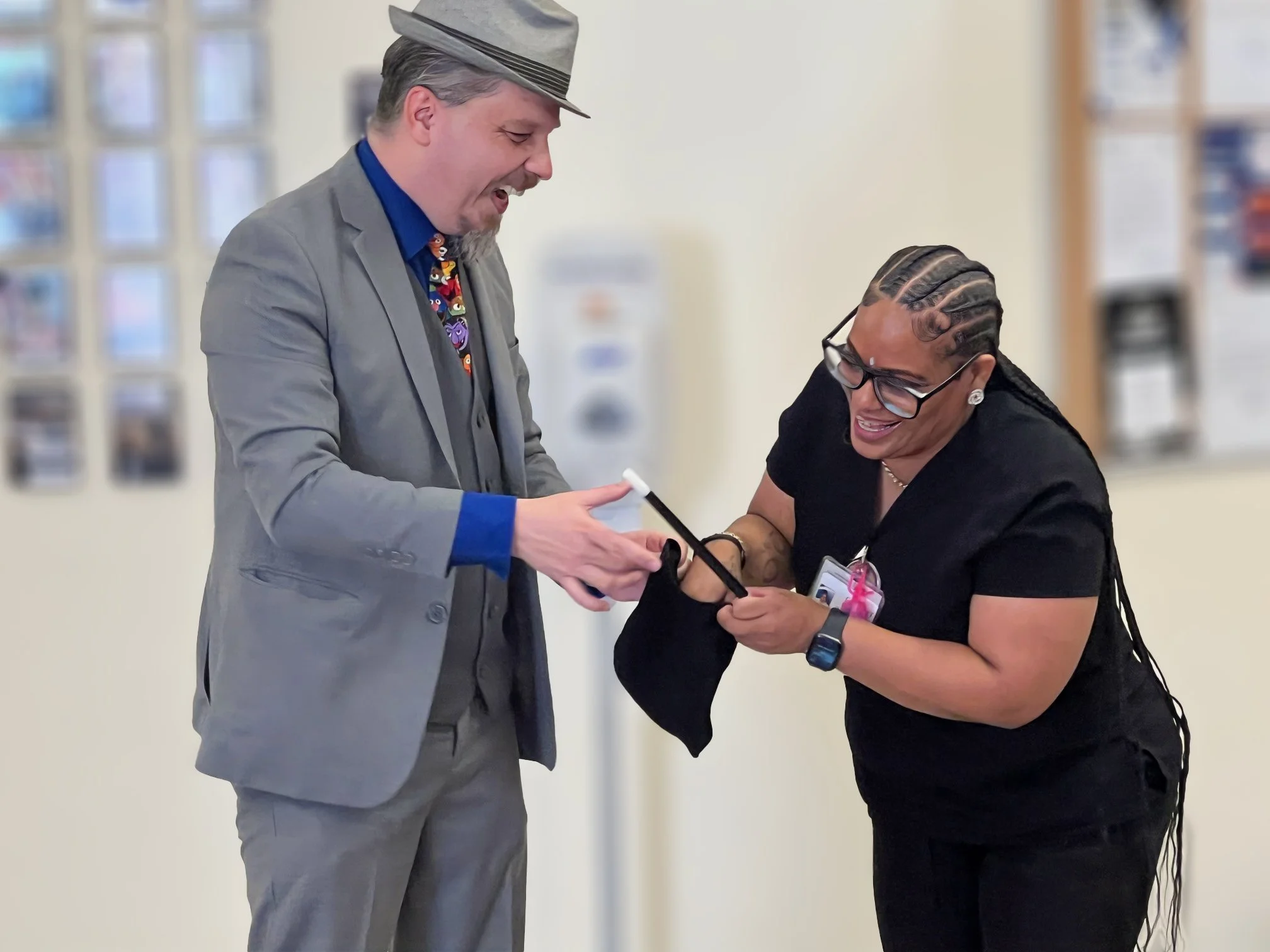 A nurse from the VA in Barstow peers in a black bag with a wide smile as John is dressed in a grey suit with a muppet tie and grey fedora smiles with her
