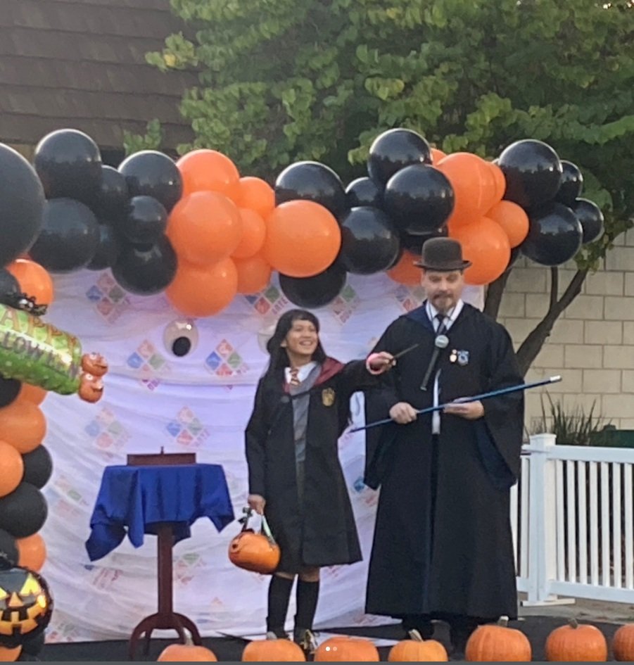 A young girl in a graduation gown and cap holding a pumpkin-shaped bucket, smiling next to a man dressed as a wizard or magician, both standing on a decorated stage with Halloween-themed balloons and pumpkins.