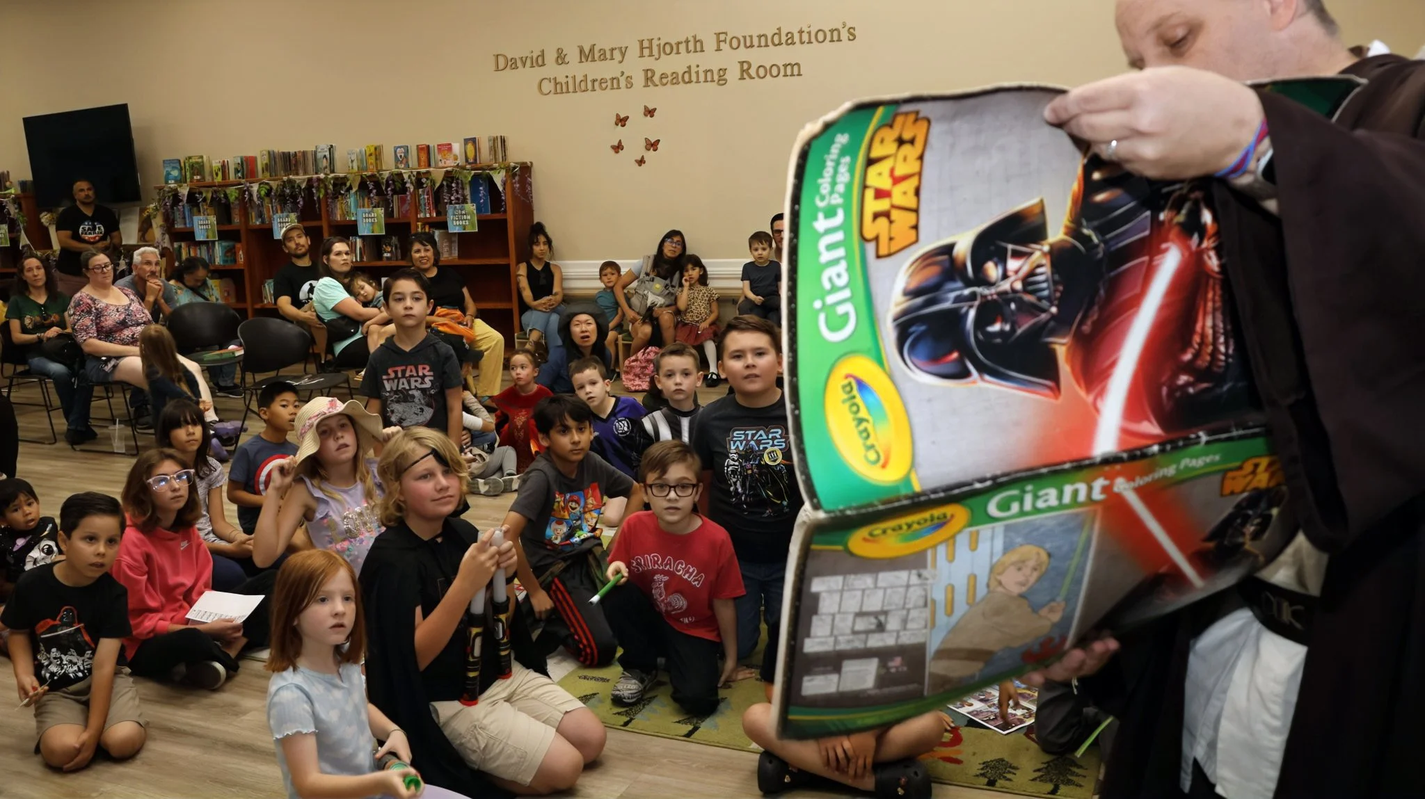 John performing the Star Wars Magic coloring book as Obi John Kenobi at the Upland library, many children look at the open coloring book, locked in to the performance. 