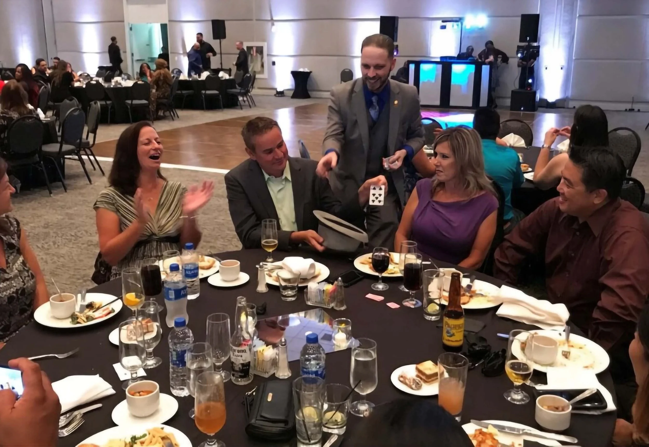 John performing for the leadership table at the Frito Lay Pepsi Corporate event. A woman gasps clapping her hands as a man holds a hat producing a card from it, several onlookers smile. 