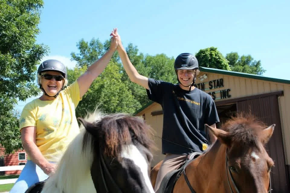 Two smiling people in helmets high-five each other while riding horses outdoors on a sunny day.
