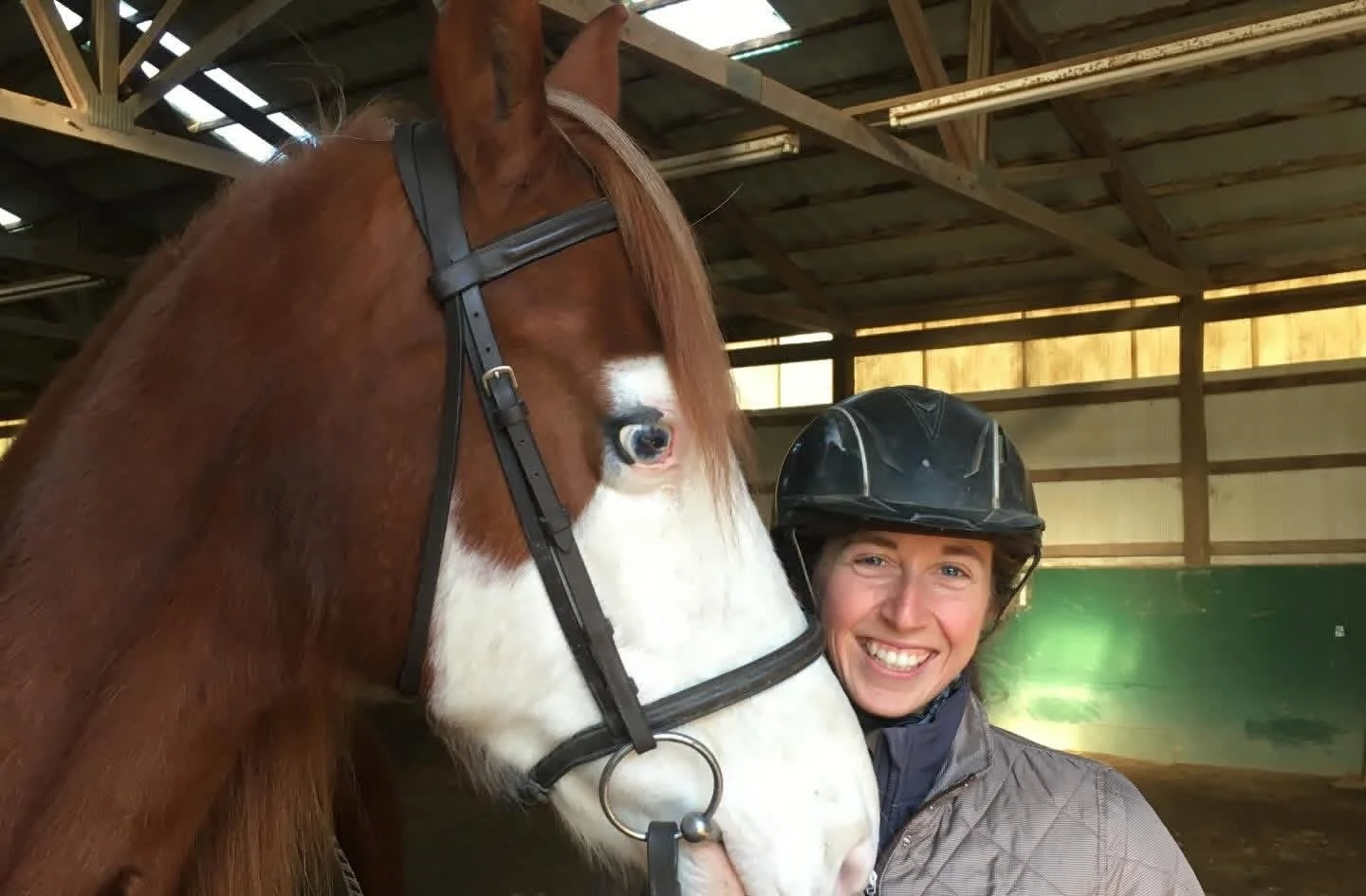 A woman in a riding helmet smiling next to a brown and white horse inside an arena.