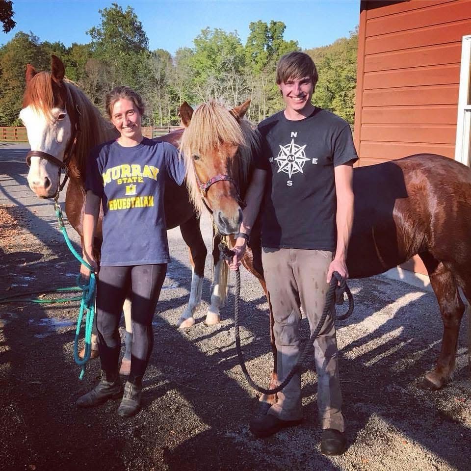 Two smiling teenagers, a girl and a boy, standing outdoors with two horses on sunny day, near a red barn and trees in the background.