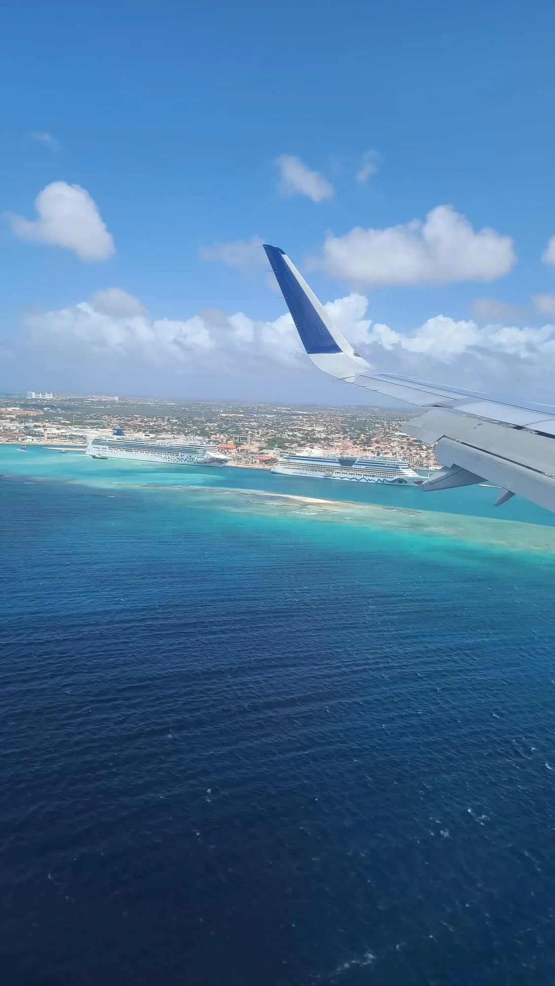 View from an airplane window showing the plane's wing, turquoise ocean, beach, and cruise ships docked at a port with a city in the background.