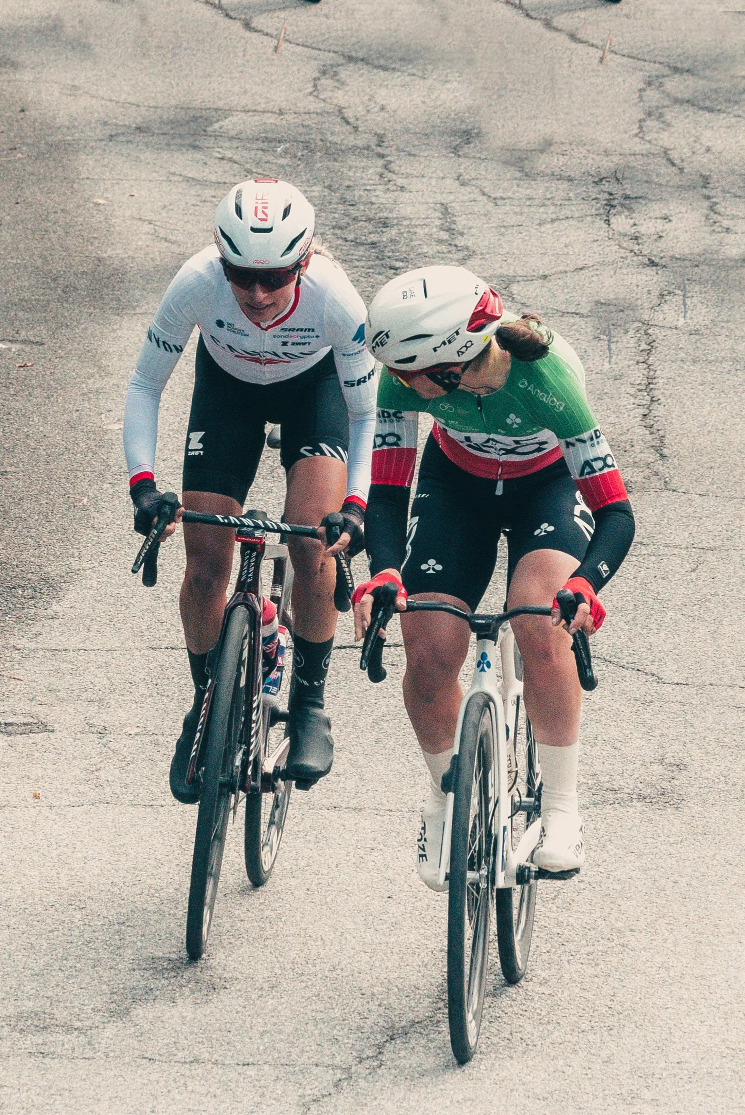 Two women riding bicycles on a cracked pavement, wearing helmets and sportswear, engaged in a conversation.