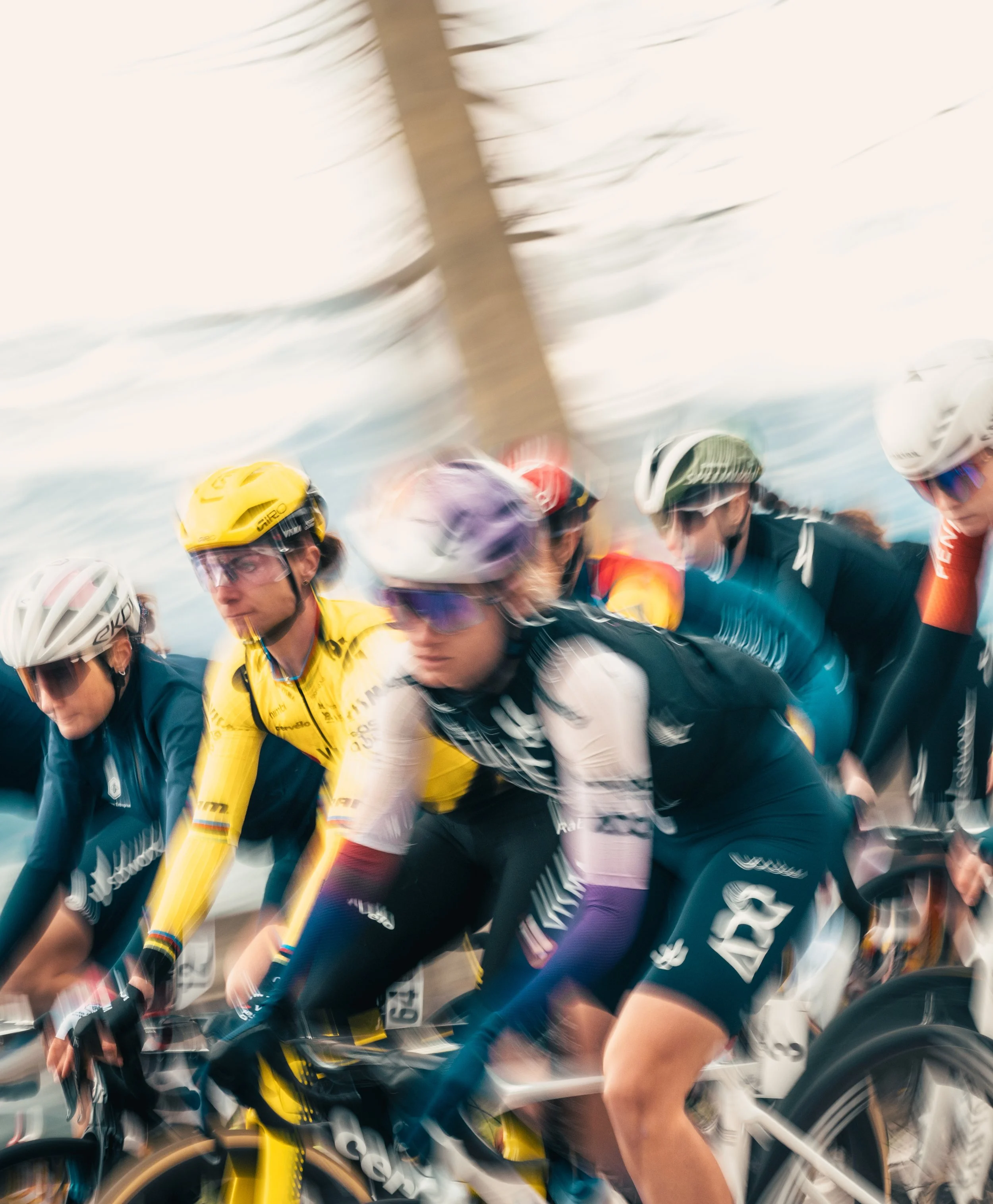 Cyclists at the start of a race, wearing helmets and colorful jerseys, with motion blur indicating high speed.