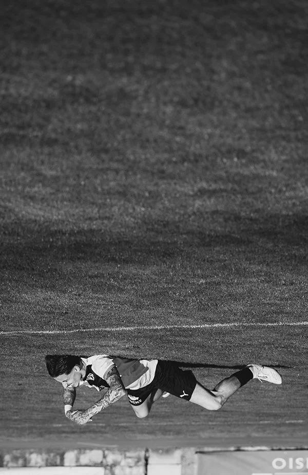 Black and white photo of a male soccer player falling on the grass field, wearing jersey, shorts, and cleats.