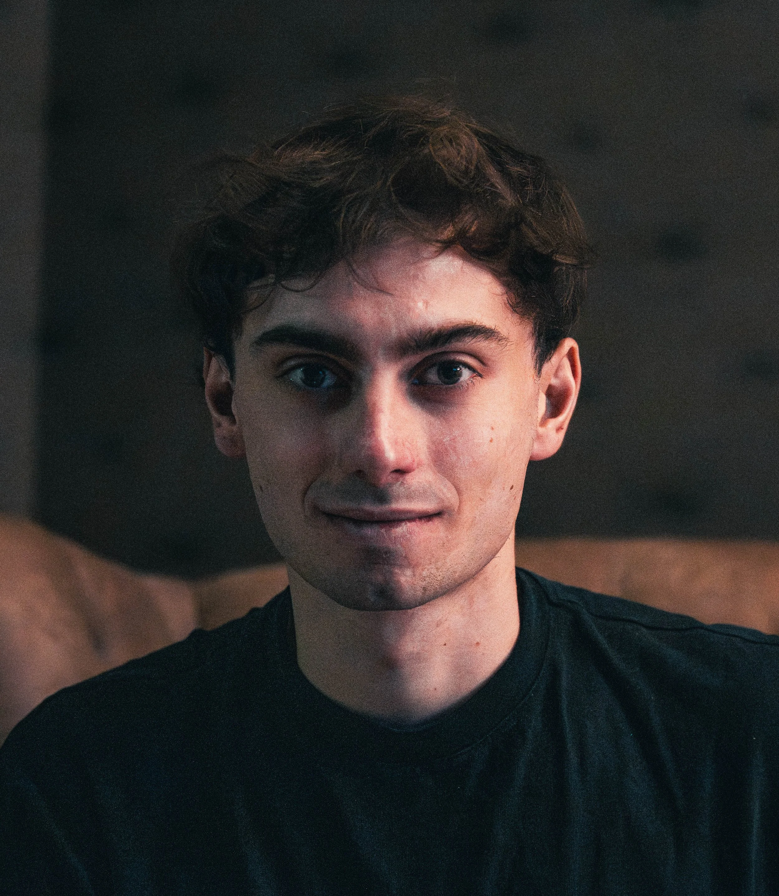 Close-up of a young person with short, tousled dark hair and fair skin, wearing a black shirt, against a dark background.