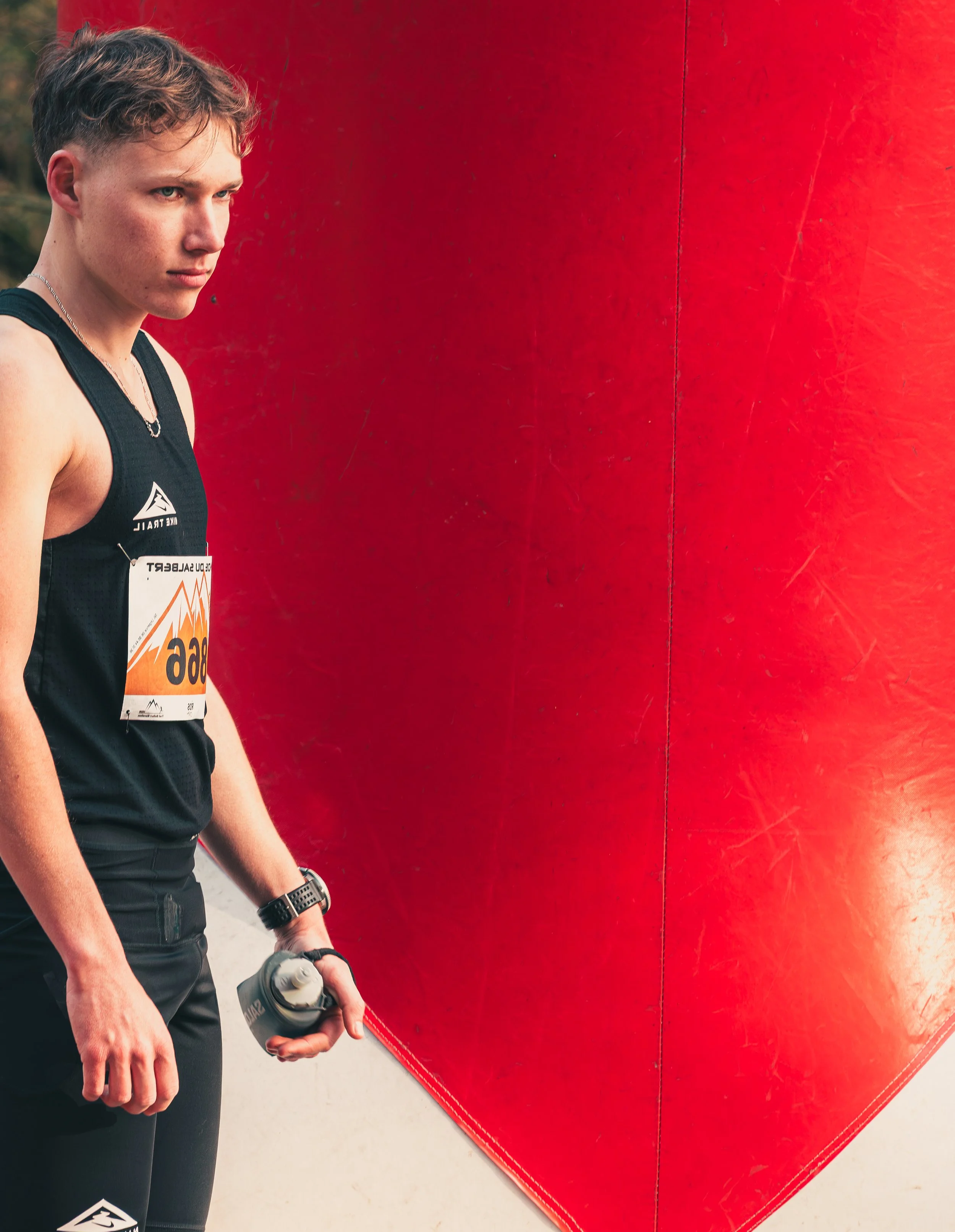 A young athlete holding a water bottle, dressed in athletic gear, standing near a large red structure, possibly a climbing or sports wall.