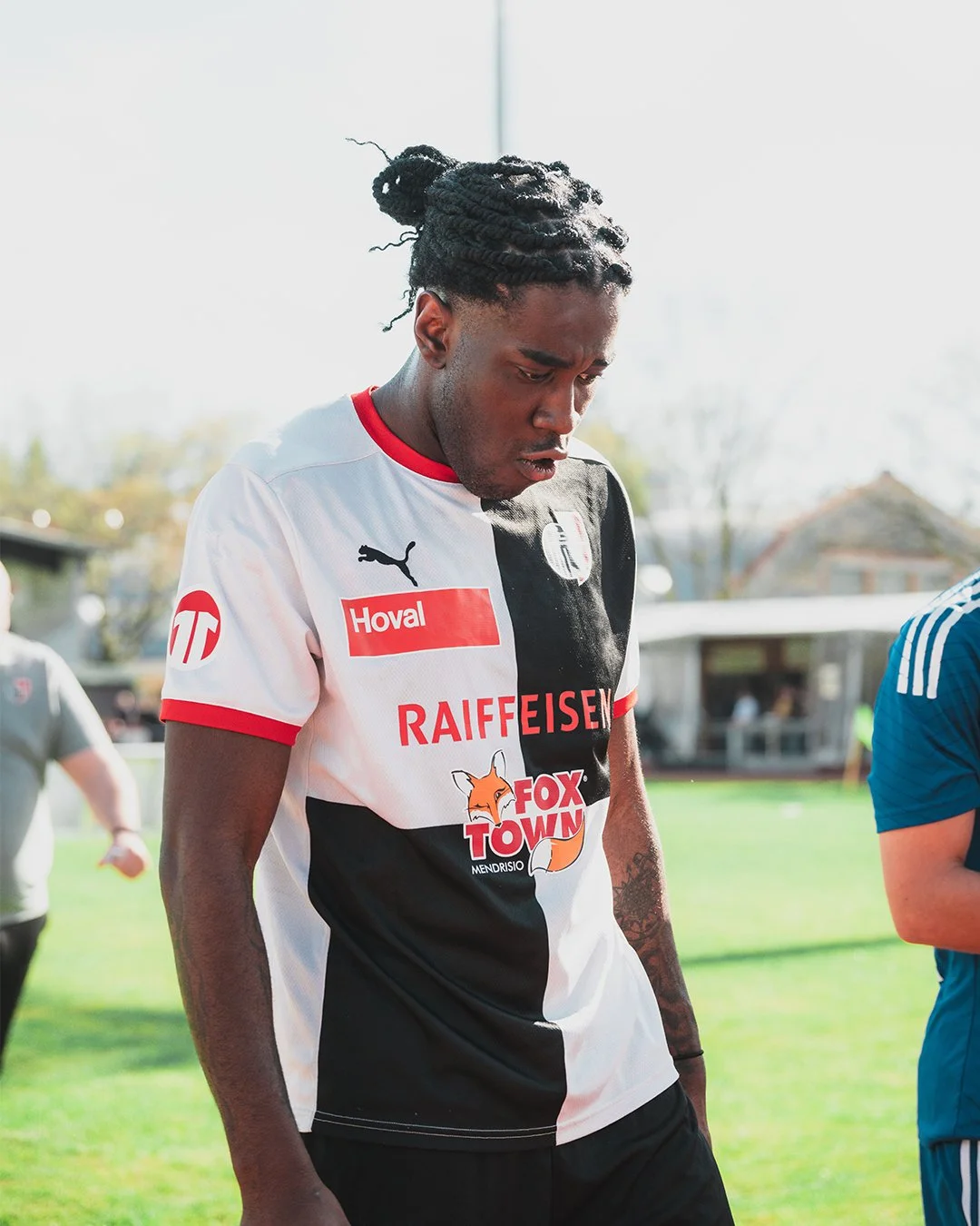 A young male soccer player in a black and white jersey with sponsor logos, looking down with a serious expression, standing on a field.