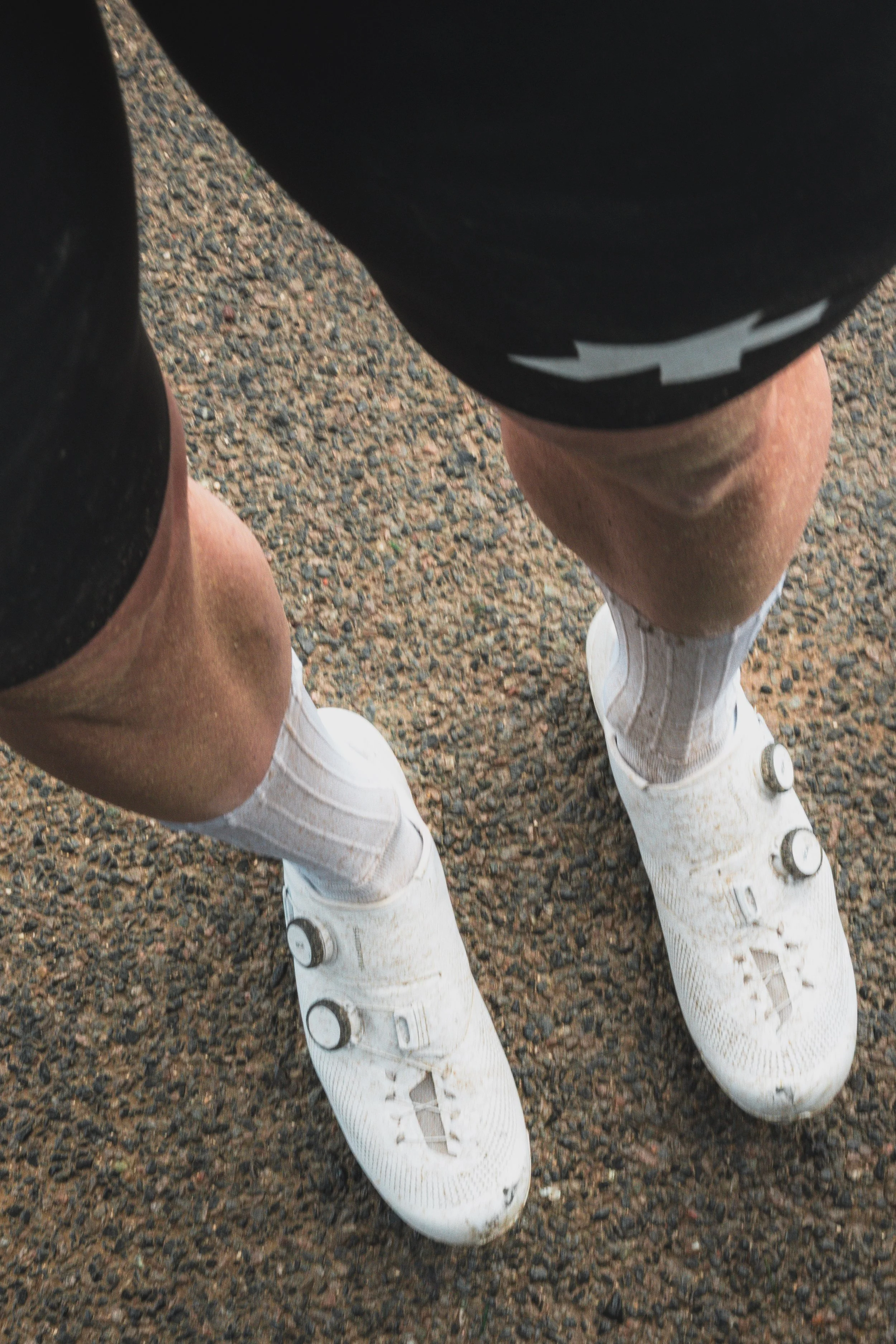 Person wearing white rollerblades, black shorts, and a black shirt, standing on a gravel path.