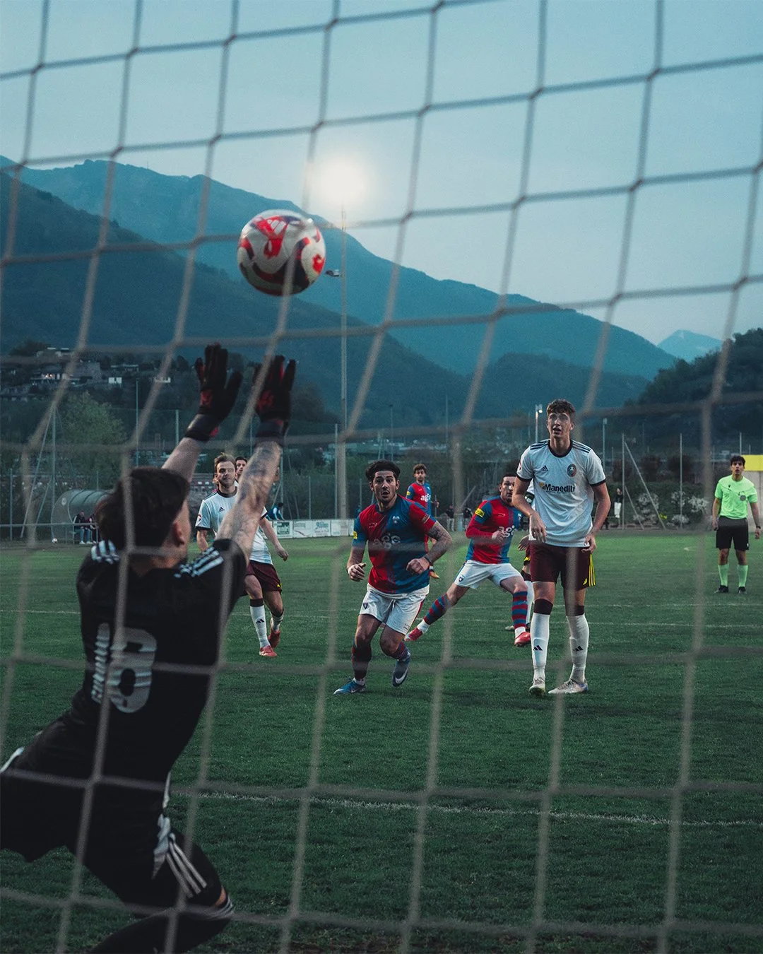 Soccer match in progress viewed from the goal, with players jumping for the ball, mountain backdrop, and cloudy sky.