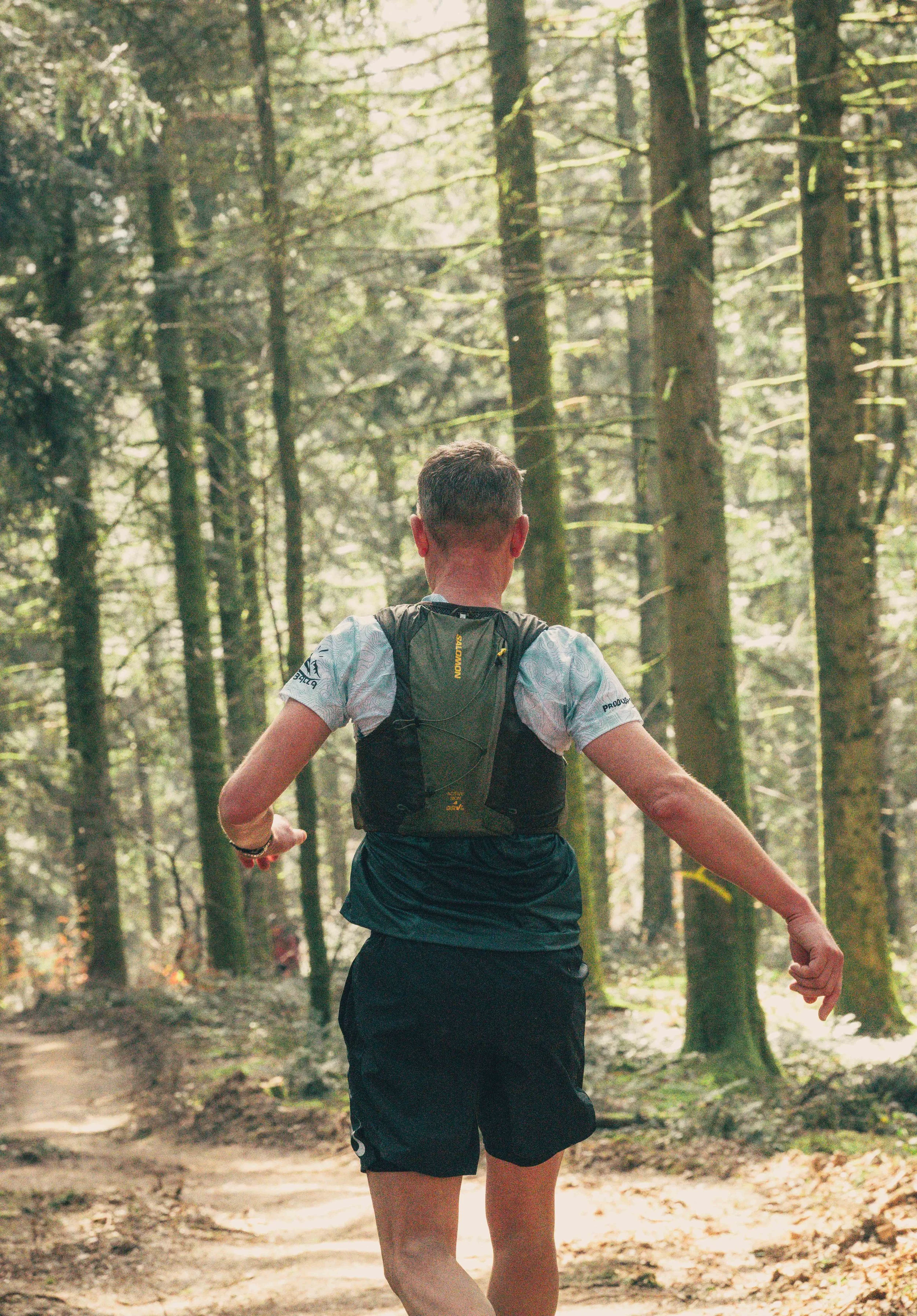 A person running or jogging through a forested trail, seen from behind, wearing a black backpack, black shorts, and a white shirt with a pattern.