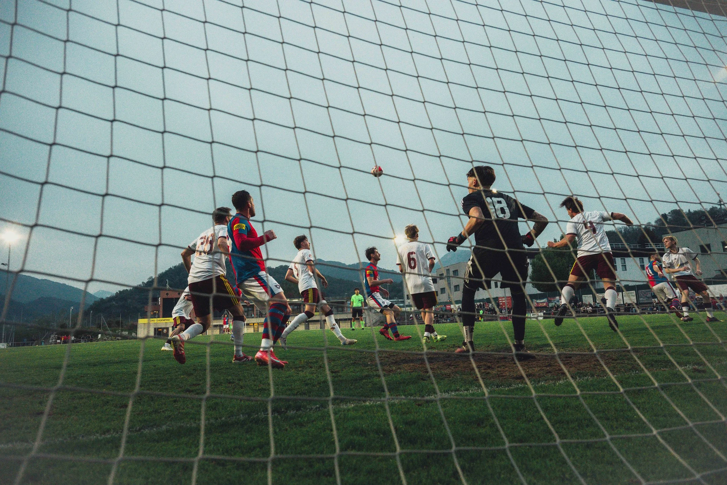 A soccer match is taking place on a field, viewed through a goal net. Players are actively moving, with some running and others preparing for the ball. The scene is set in a stadium with mountains and buildings in the background, under a cloudy sky.