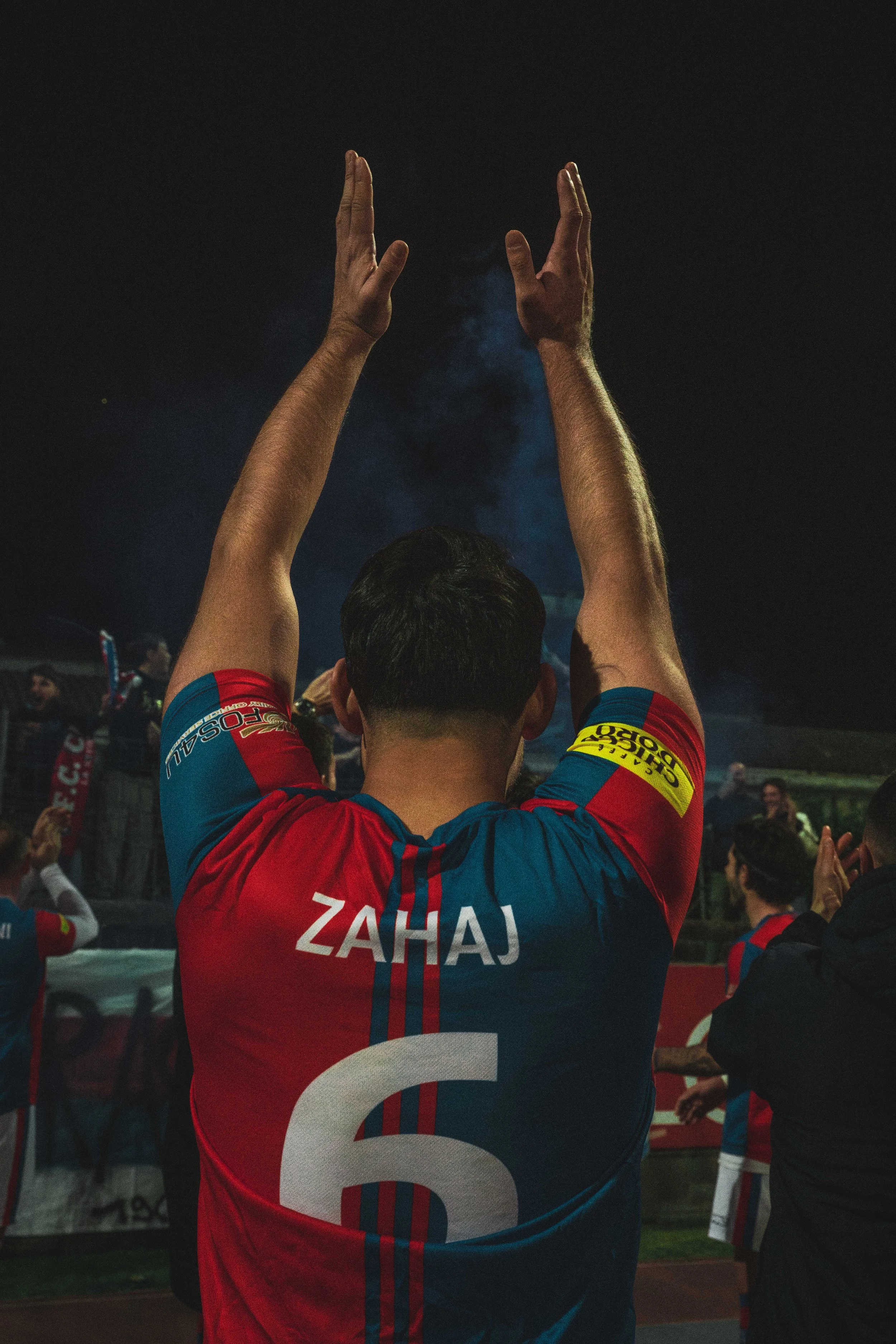 A person wearing a red, blue, and white sports jersey with the name 'Zahaj' on the back, raising both hands above their head in a stadium or sports event at night, with other people and banners visible in the background.