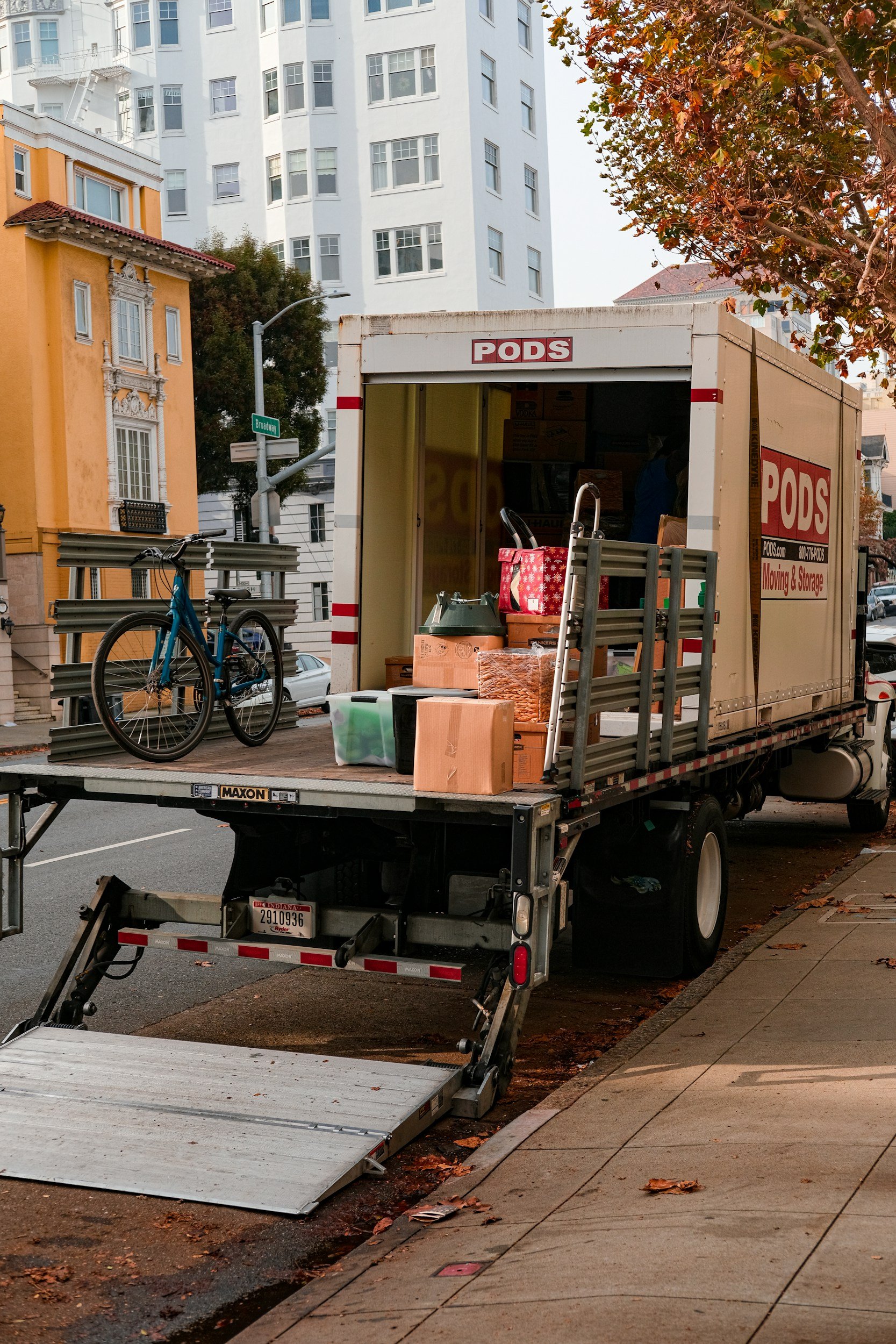 Moving truck parked on city street with boxes and a bicycle on the back, surrounded by residential buildings and trees with fall foliage.