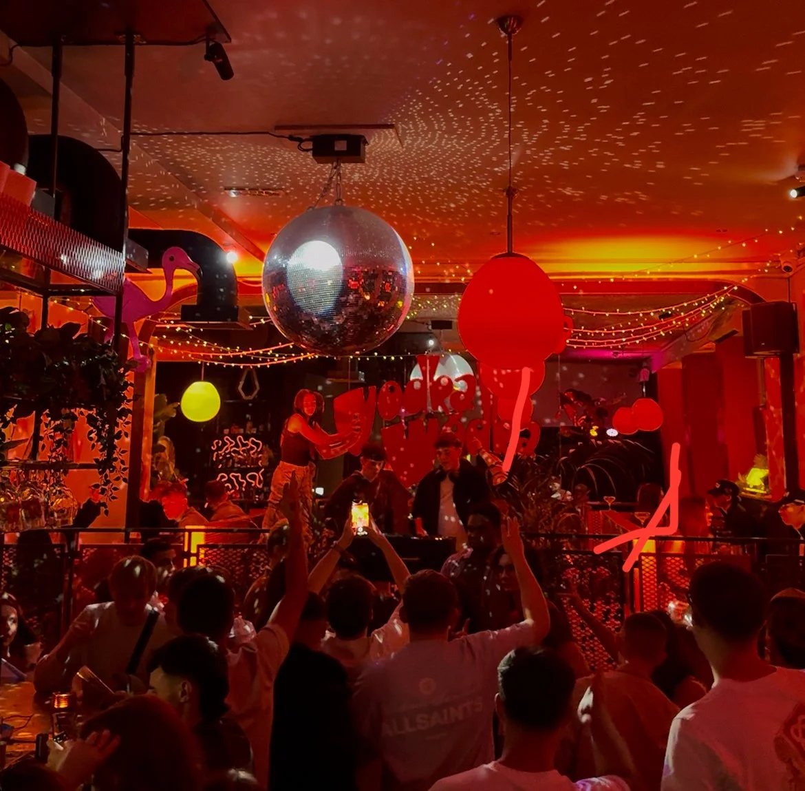 A lively indoor party scene with a disco ball hanging from the ceiling, colorful hanging lights, and people dancing and enjoying themselves. The room is decorated with neon and string lights, with a woman standing on a table or platform, possibly a DJ or performer, surrounded by a crowd.