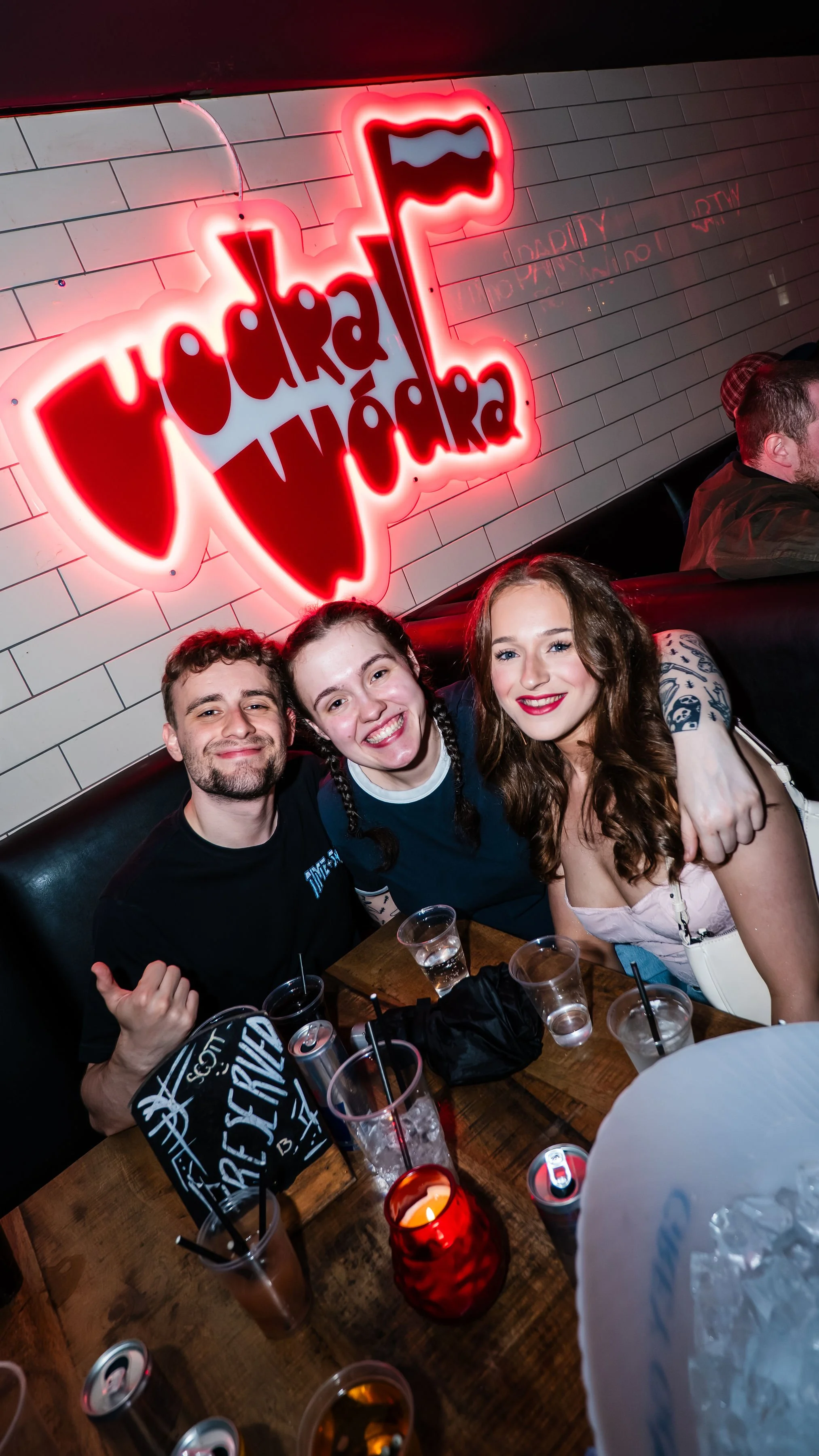 Three friends sitting together at a table in a bar or restaurant, smiling. The background has a neon sign with a red outline and text, white tiled wall, and other patrons. The table has drinks, cans, and a ballot box decorated with graffiti.