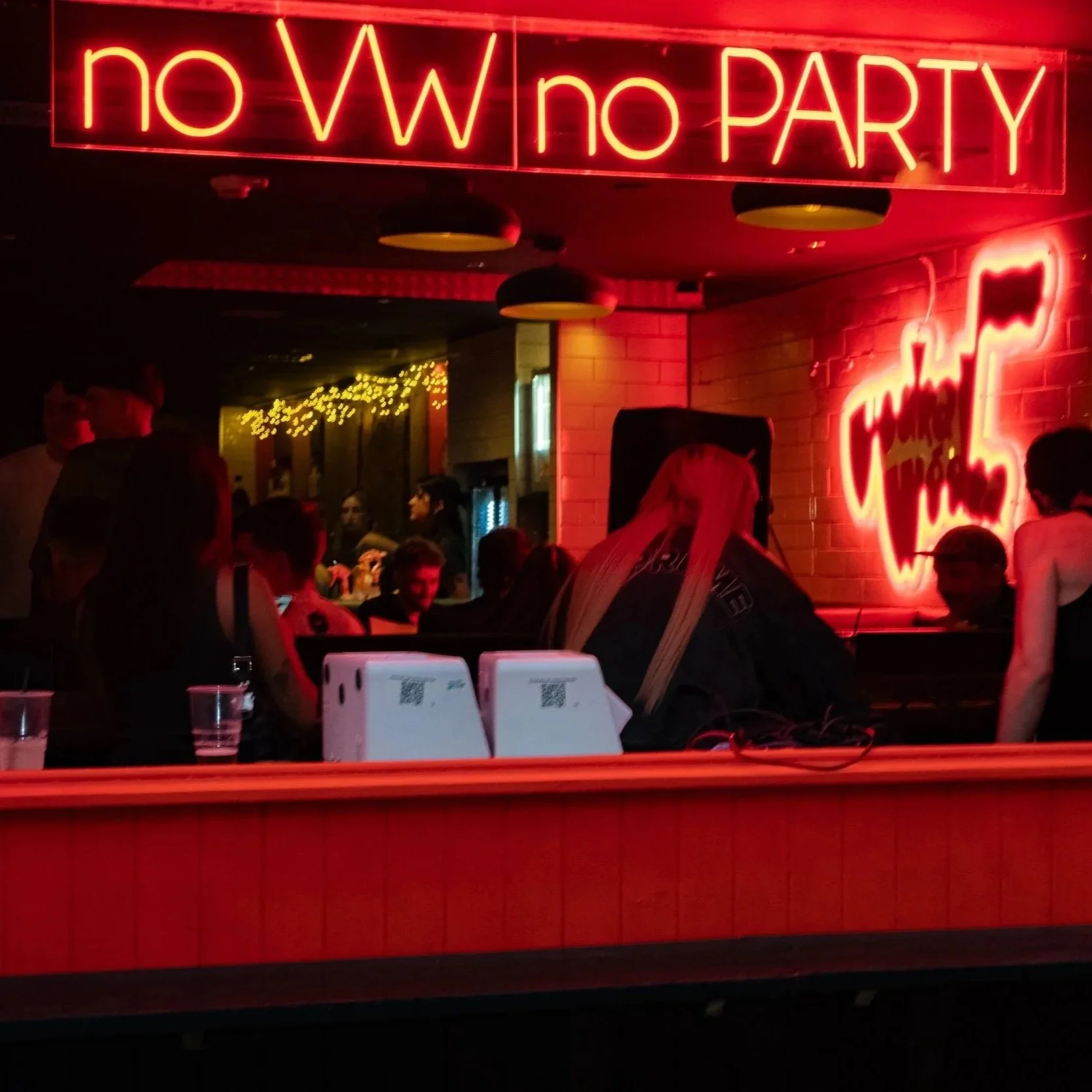 A neon sign in red with the message 'no W no PARTY', and another neon sign showing 'special' in on a brick wall. There are people gathered in a dimly lit venue with string lights, tables, and drinks, suggesting a party or social gathering.