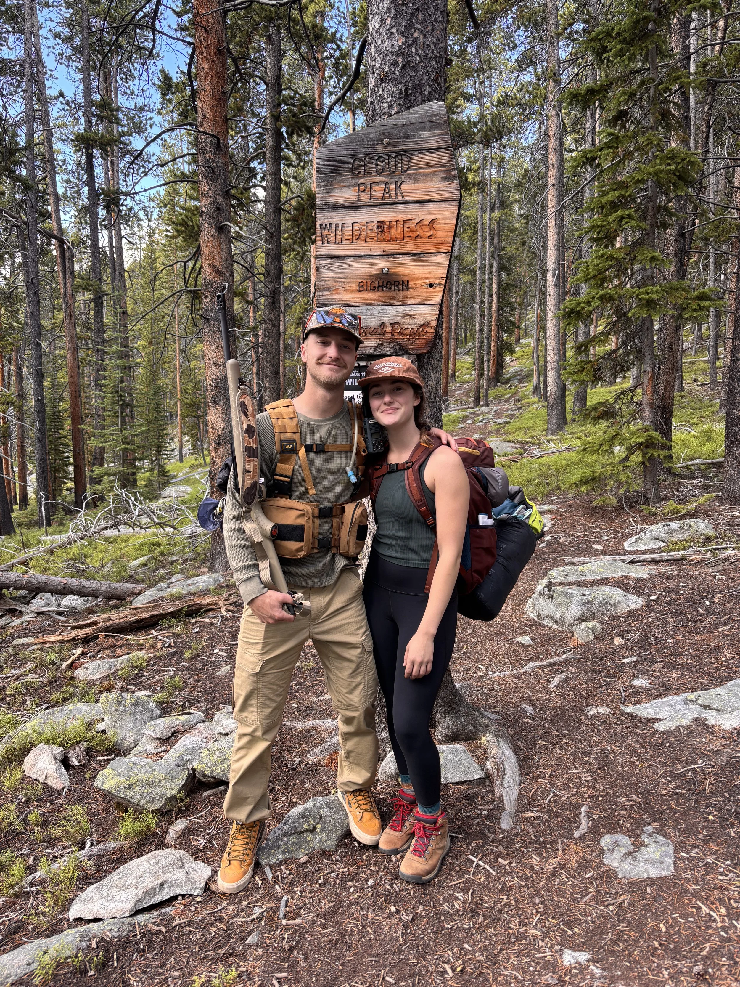 A man and woman in hiking gear standing next to a wooden sign in a forest, with trees and rocks in the background.
