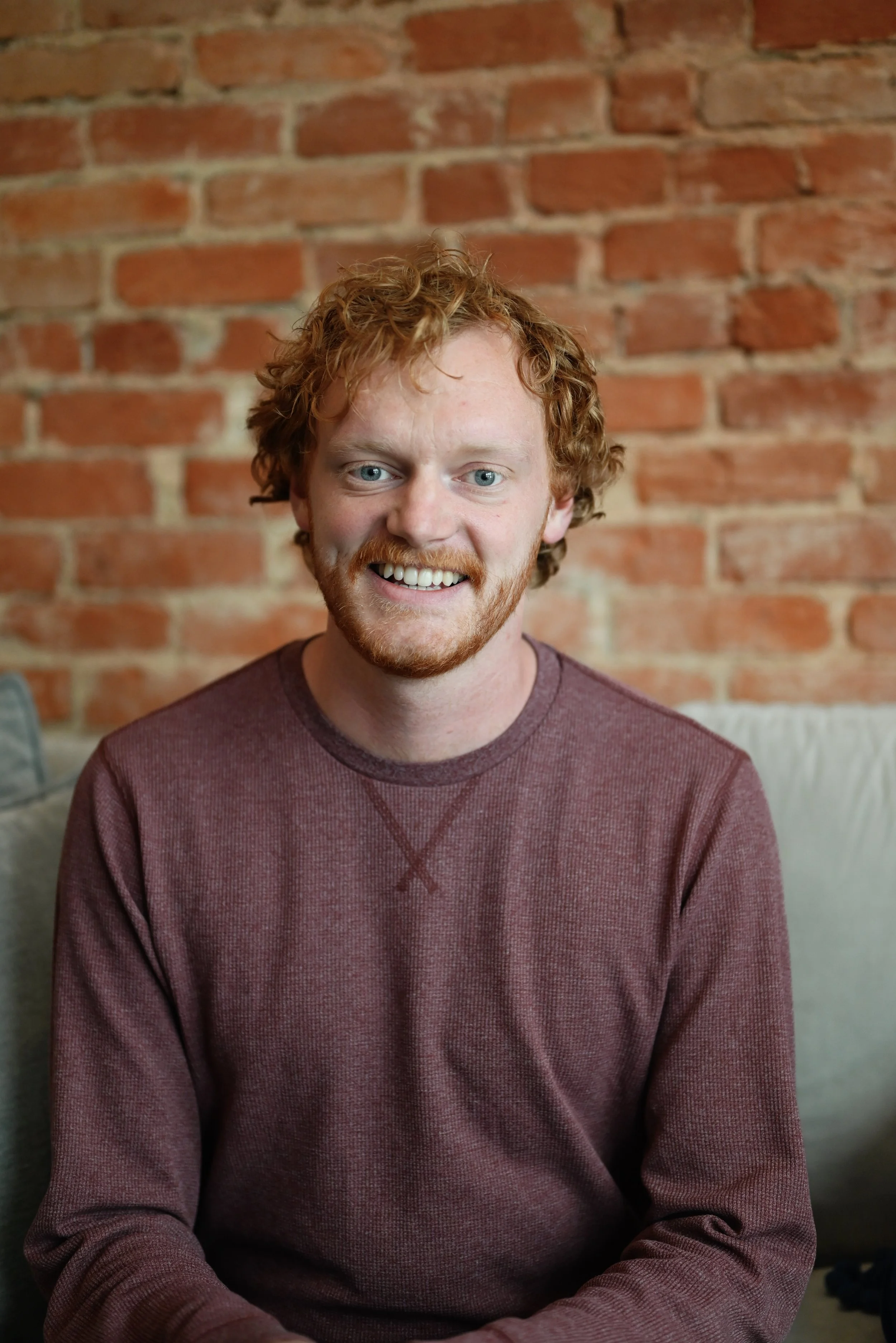 Young man with curly red hair, beard, and blue eyes, smiling, wearing a maroon long-sleeve shirt, sitting in front of a brick wall.