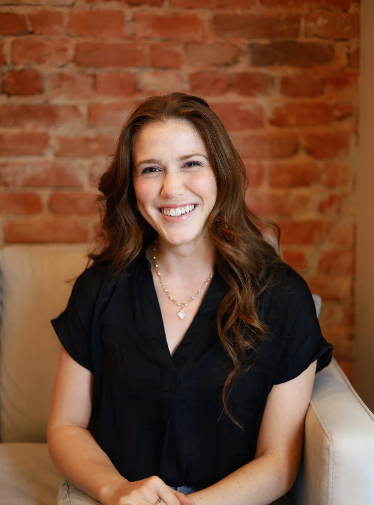 A woman with long, wavy brown hair, smiling, sitting on a beige sofa, wearing a black top and a pearl necklace, in front of a brick wall.