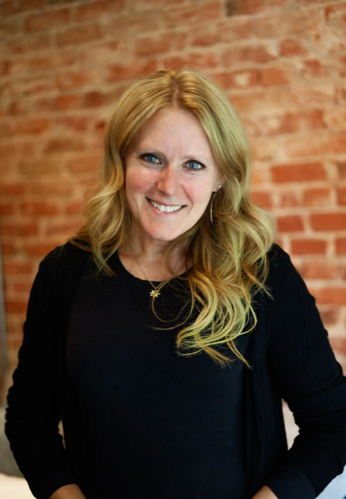 A woman with long, wavy blonde hair, blue eyes, and wearing a black top, standing in front of a red brick wall, smiling.