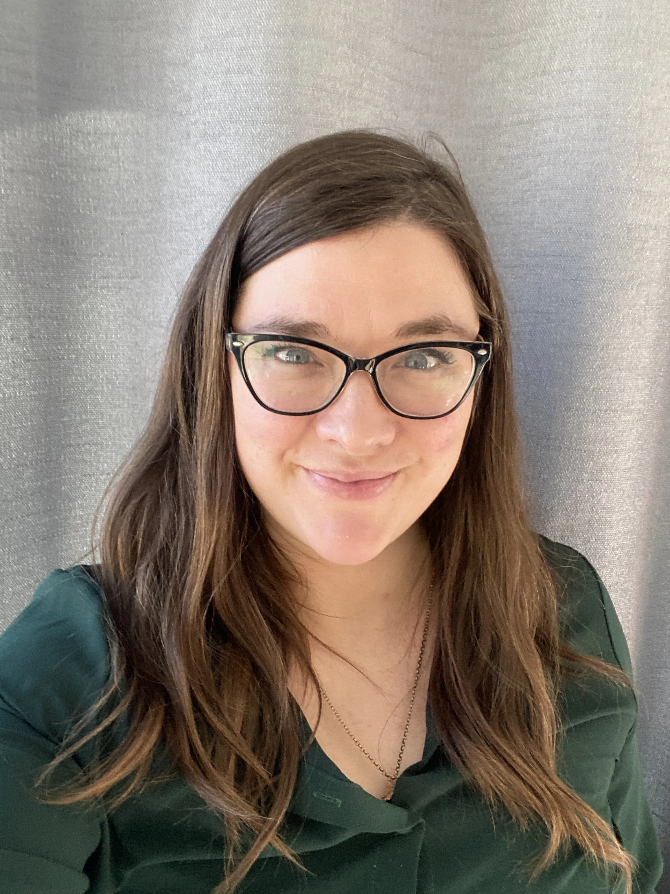 Close-up of a young woman with long brown hair, glasses, and a smile, wearing a dark green top, against a grayish background.