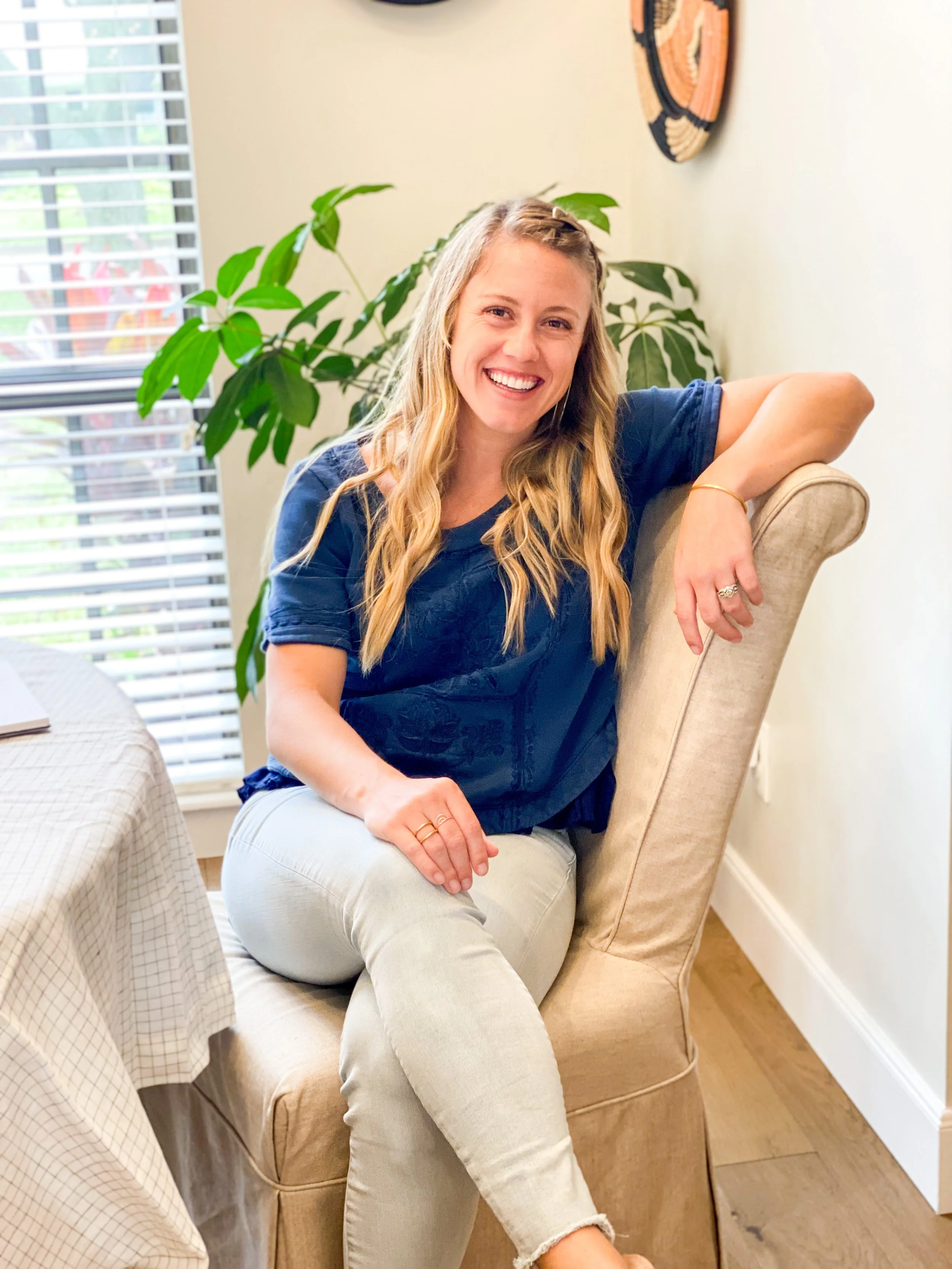 A smiling woman with long wavy blonde hair sitting in a beige armchair near a large window with blinds, a green plant behind her, wearing a navy blue top and light-colored pants.