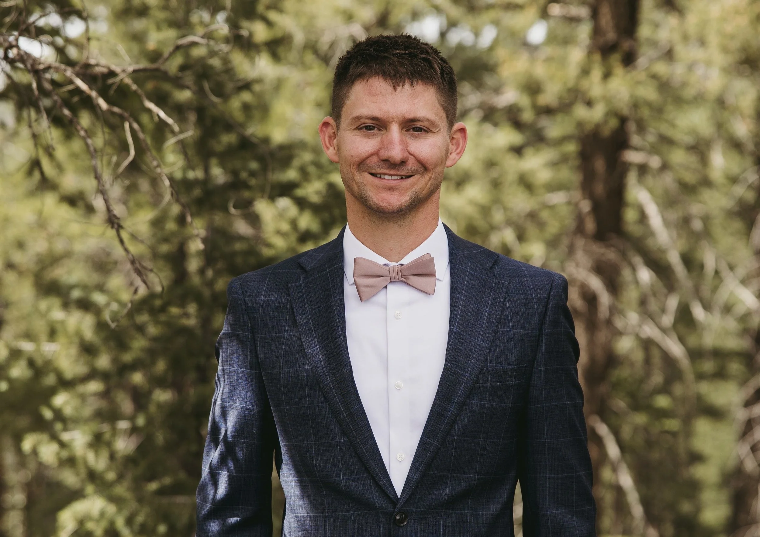 A man in a suit with a light pink bow tie standing outdoors in front of trees.