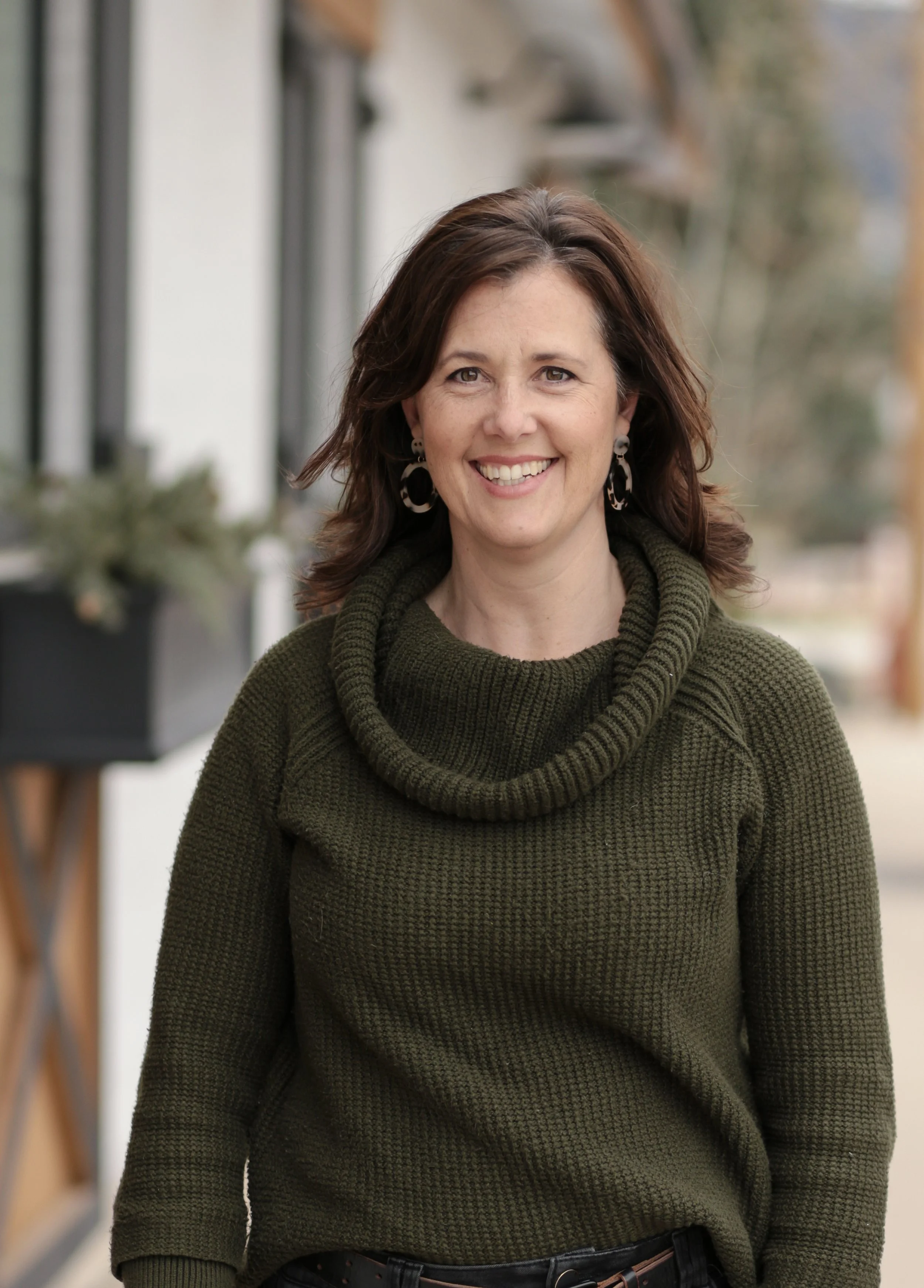 A woman with shoulder-length brown hair, wearing a dark green knit sweater with a cowl neck, standing outdoors with a blurred background of buildings and trees.