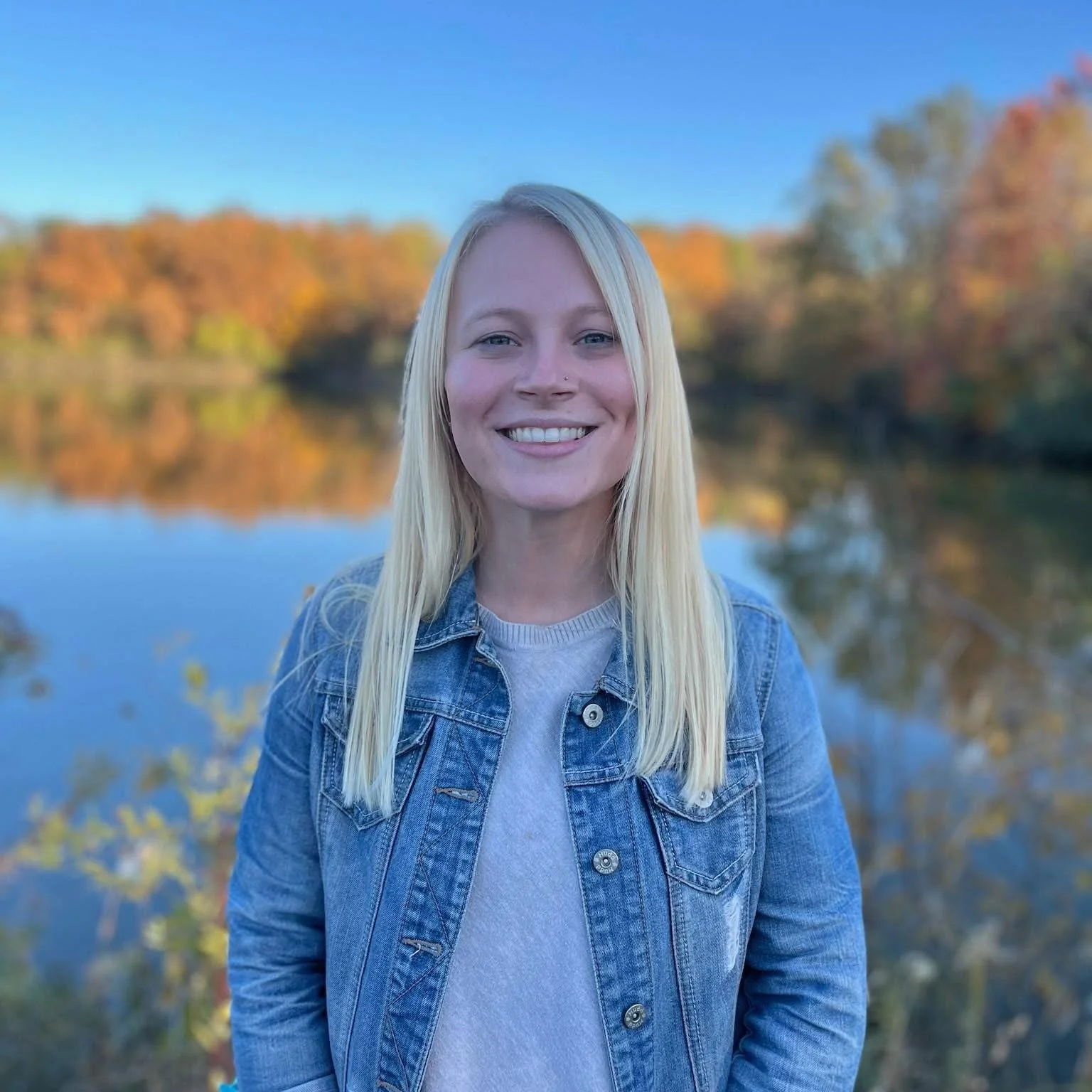 A smiling woman with long blond hair, wearing a denim jacket over a gray t-shirt, standing outdoors near a body of water with trees displaying fall colors in the background.