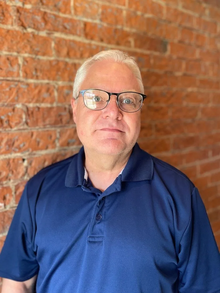 A middle-aged man with gray hair and glasses, wearing a navy blue polo shirt, standing against a brick wall.