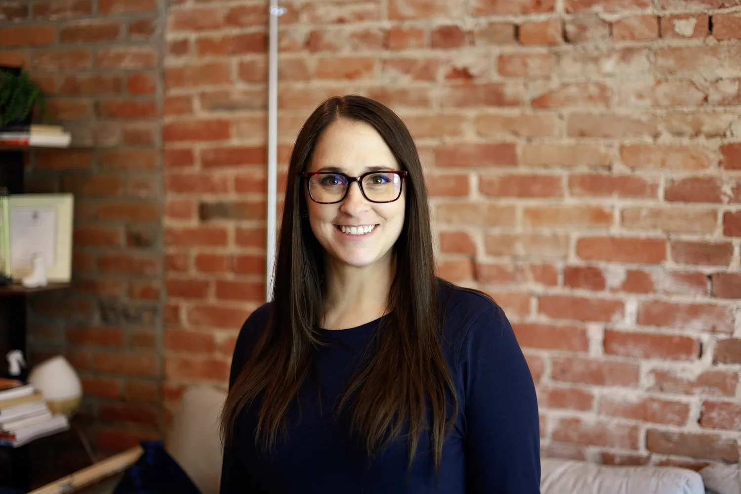 A woman with long brown hair and glasses smiling at the camera in front of a red brick wall.
