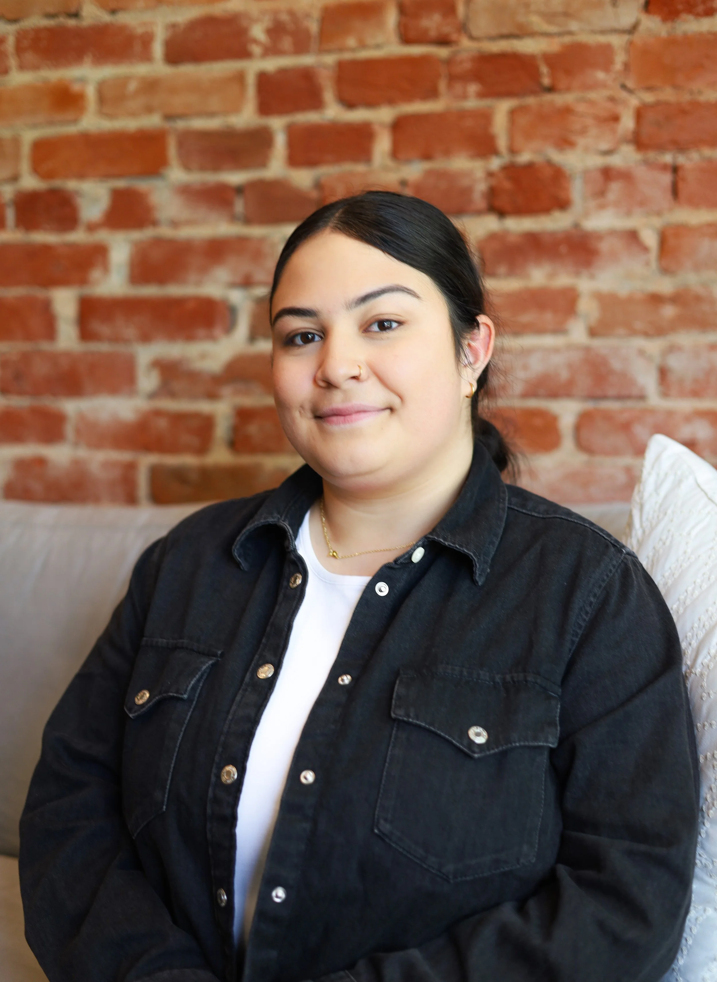 A young woman with dark hair, wearing a nose ring, gold earrings, a gold necklace, a white shirt, and a black denim jacket, sitting on a beige couch with a white cushion, in front of an exposed red brick wall.