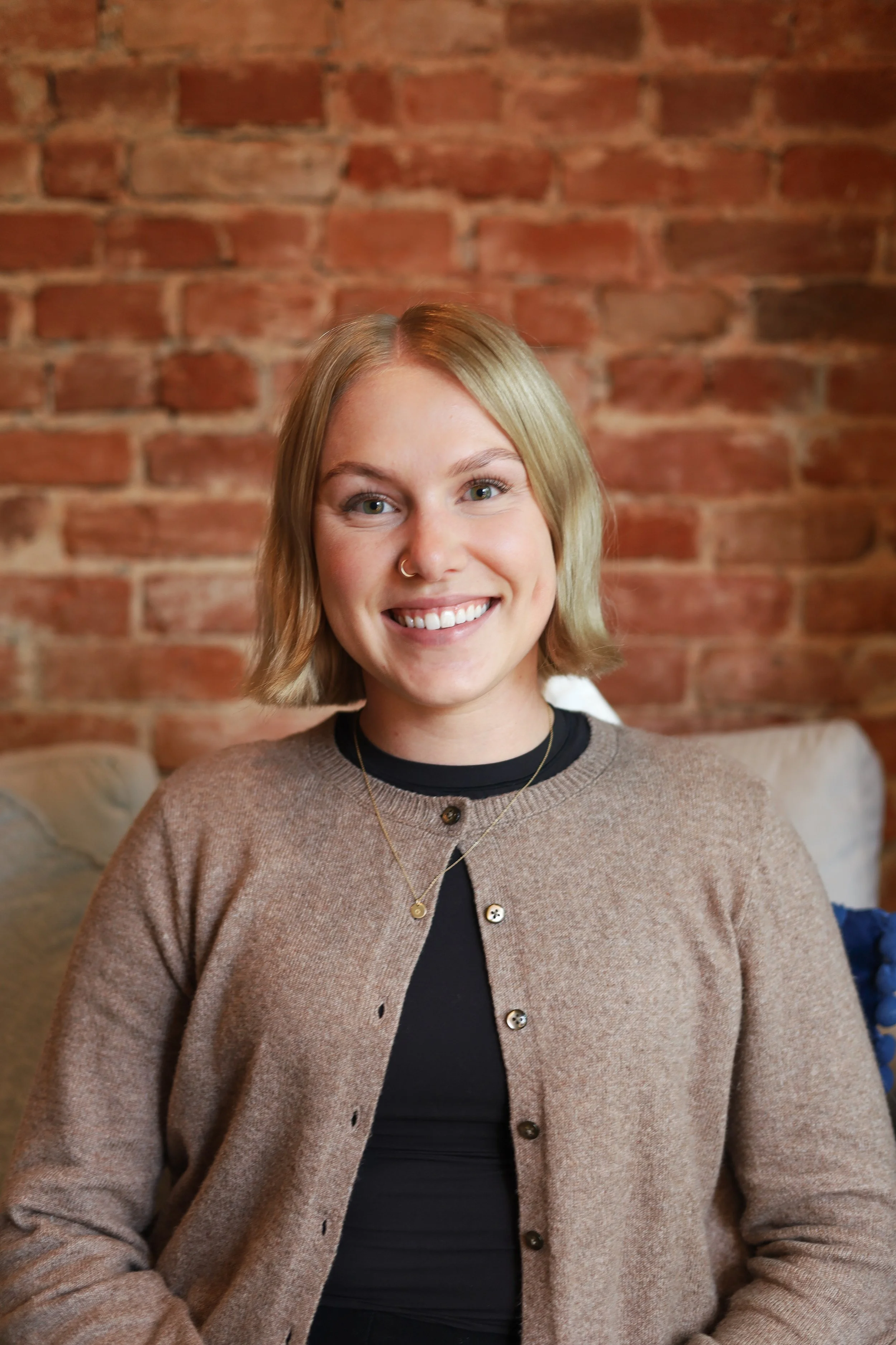 A young woman with blonde hair smiling in front of a brick wall, wearing a beige cardigan over a black top with layered necklaces.