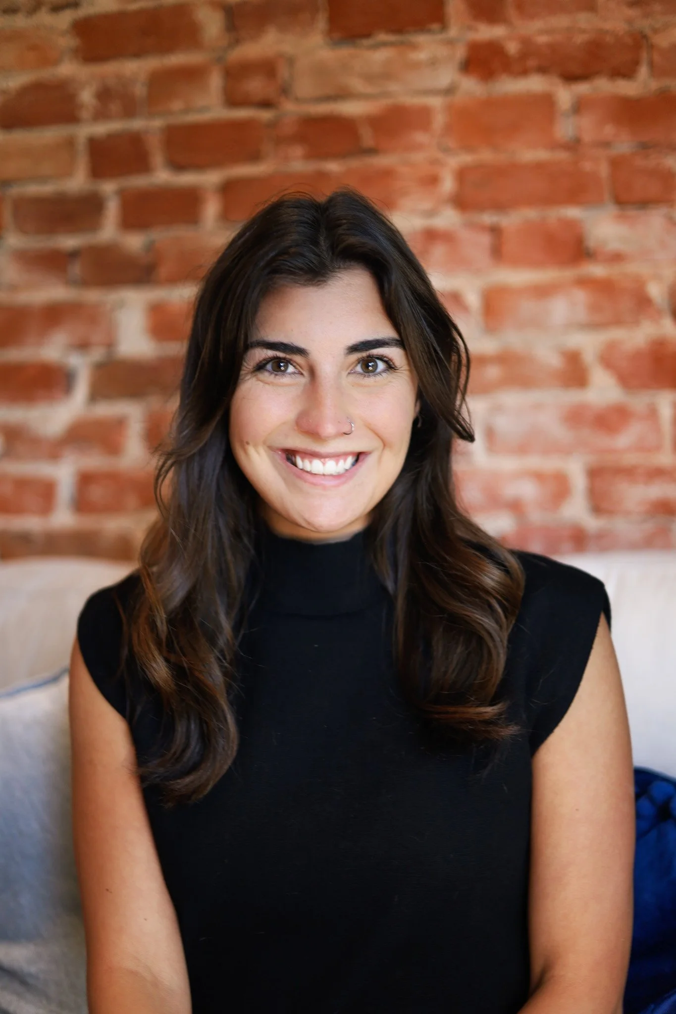 A young woman with long brown hair and a nose piercing is smiling at the camera. She is wearing a black top, and her shoulders are visible. She is sitting indoors against a brick wall background.