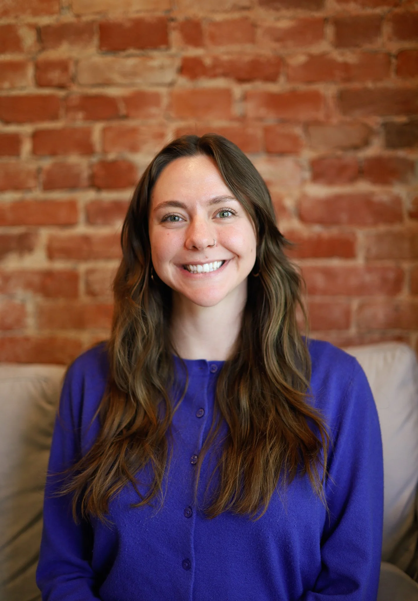 A smiling woman with long wavy brown hair and a septum piercing, wearing a royal blue button-up shirt, sitting in front of a brick wall.