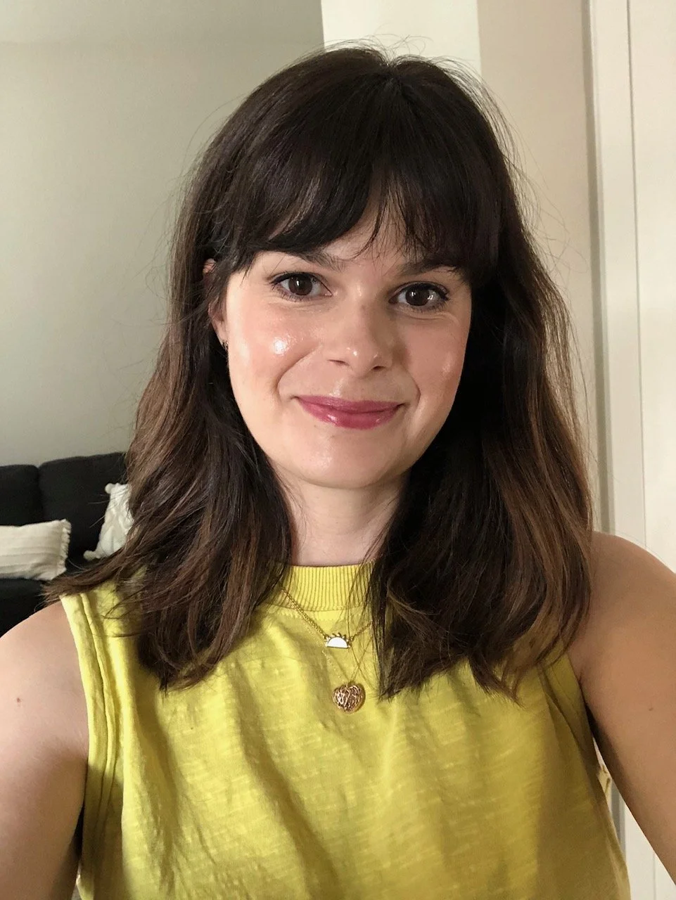 A woman with shoulder-length brown hair and bangs, wearing a sleeveless yellow top, smiling at the camera in a living room with neutral walls and black couch with pillows in the background.