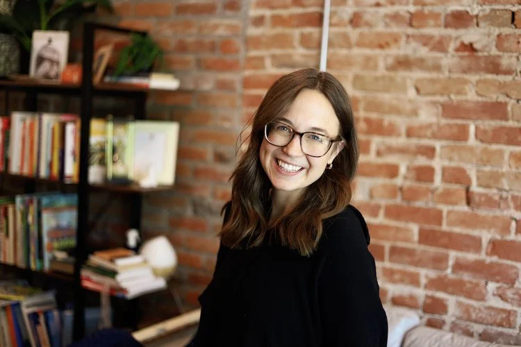 A woman with glasses and long brown hair smiling in a cozy room with a brick wall and bookshelf.