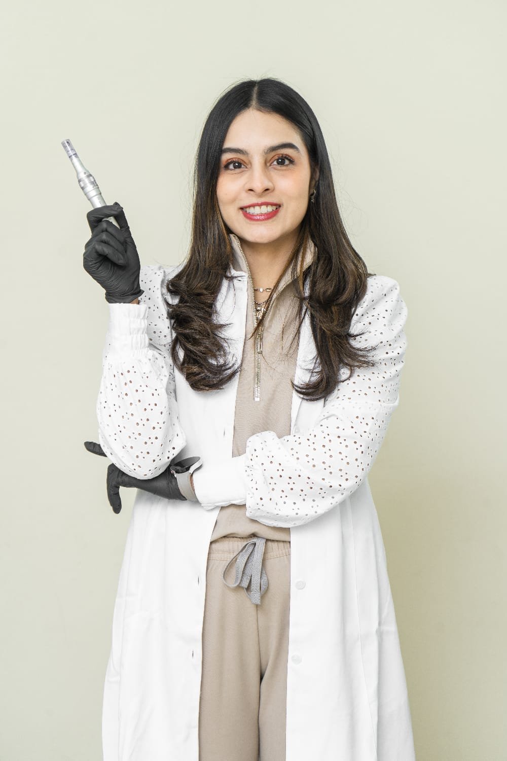 Mujer con bata blanca y guantes negros sosteniendo una punta de dispositivo dental o de tatuajes, sonriendo, frente a fondo beige.