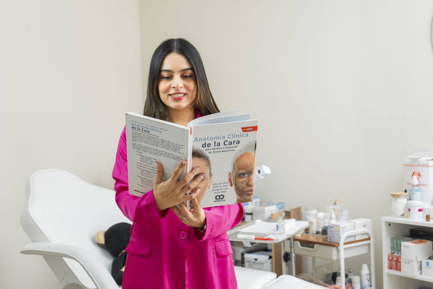 Una mujer en bata rosa revisando un libro titulado 'Anatomía Clínica de la Cara' en un consultorio médico, con instrumentos y medicinas en el fondo.